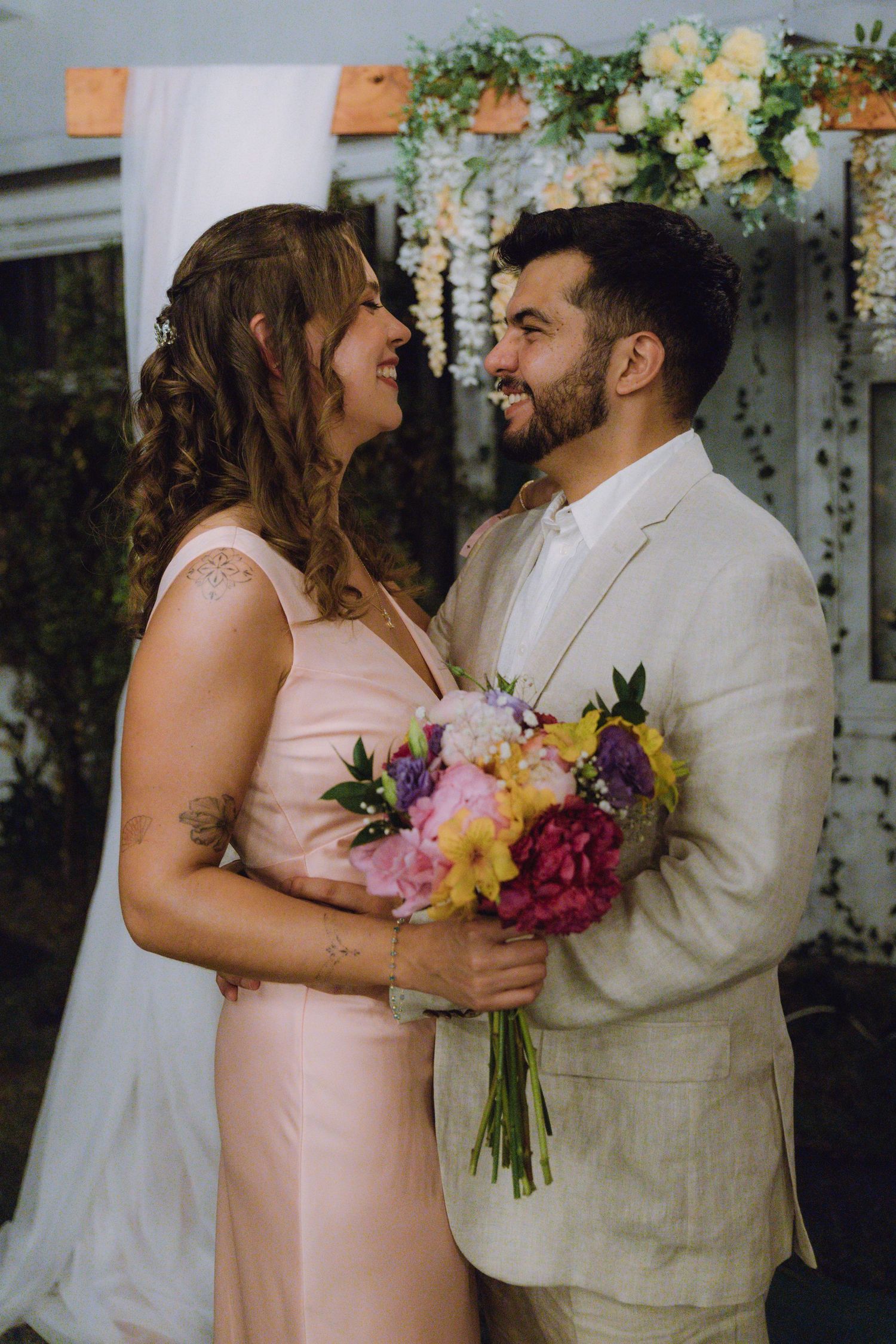 Newlyweds embrace and smile at each other under floral arch holding colorful wedding bouquet.