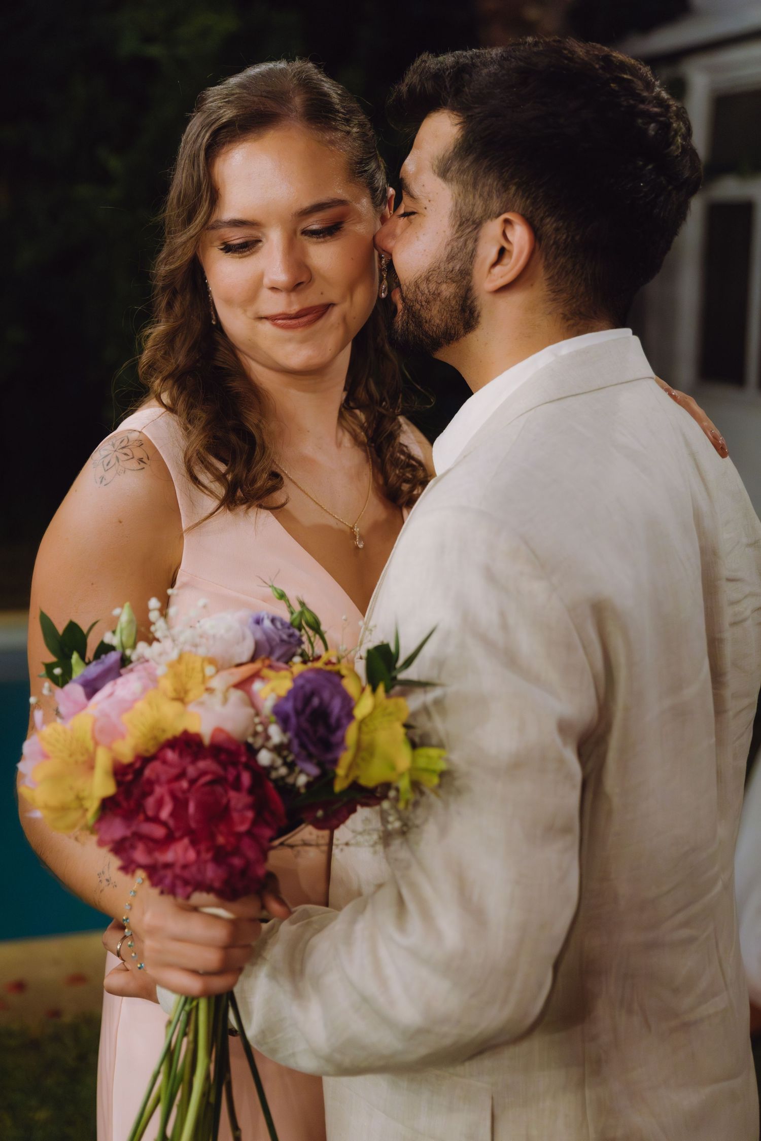 Groom kissing bride on cheek as she smiles holding colorful wedding bouquet of pink hydrangeas and yellow flowers.