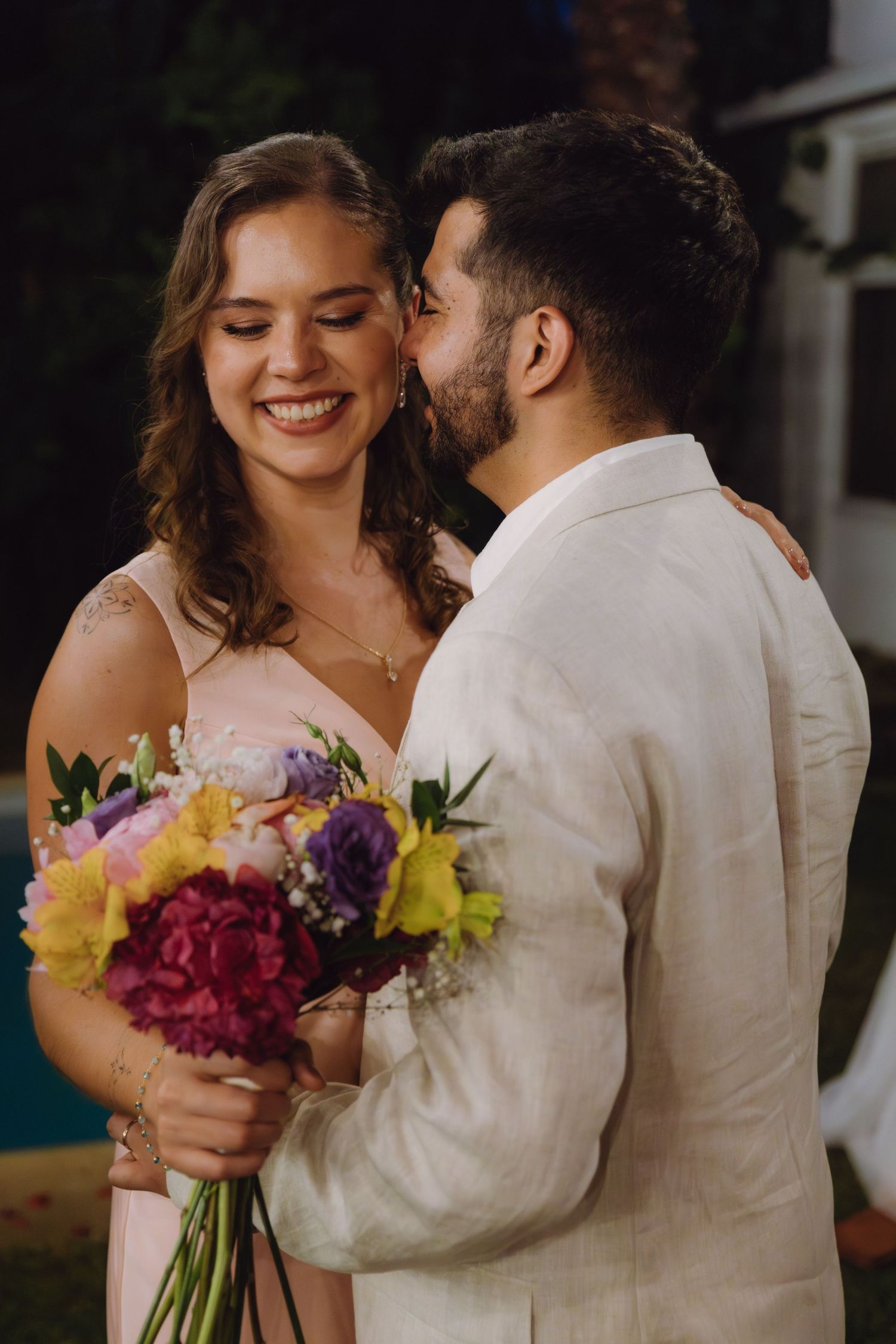 Smiling bride in pink dress holds vibrant bouquet while groom whispers in her ear at outdoor wedding.