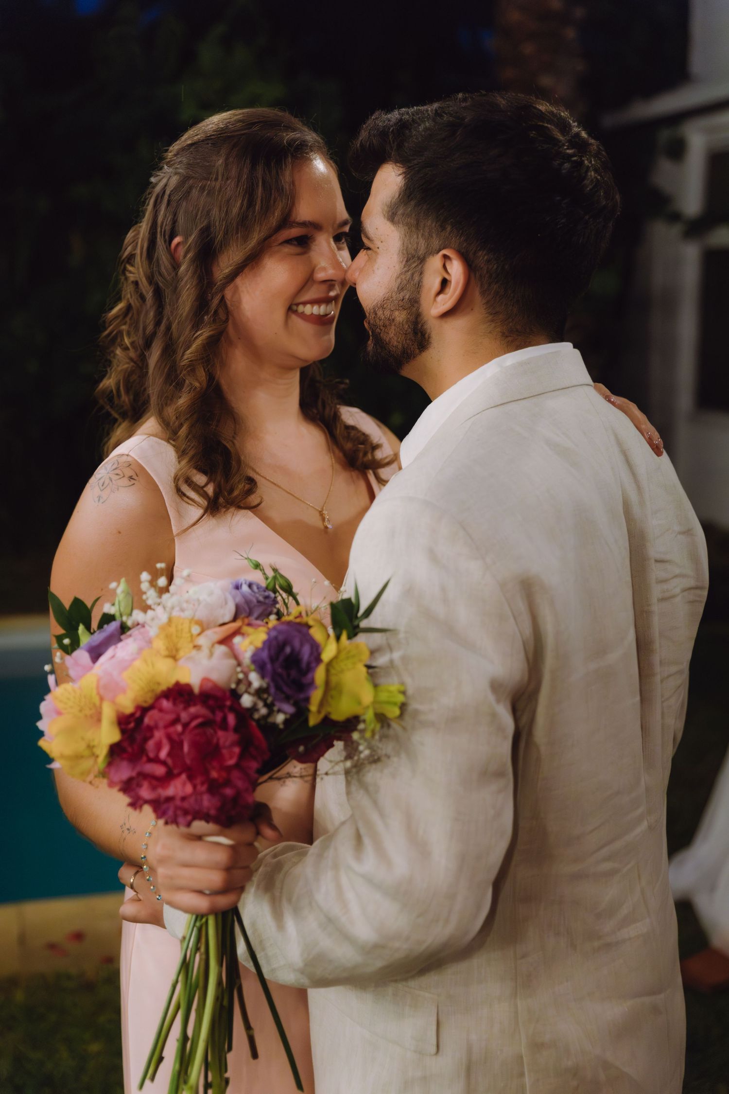 Bride and groom share a romantic nose-to-nose moment while she holds a colorful wedding bouquet outdoors.