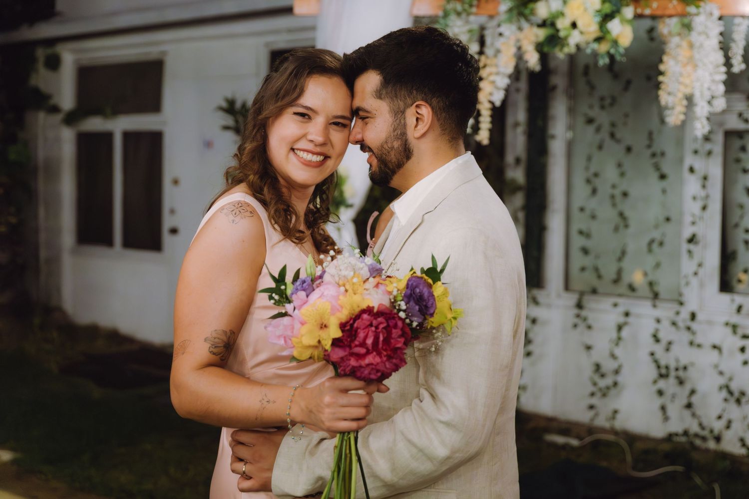Happy couple shares intimate moment touching noses, bride holding bright mixed bouquet at outdoor wedding venue.
