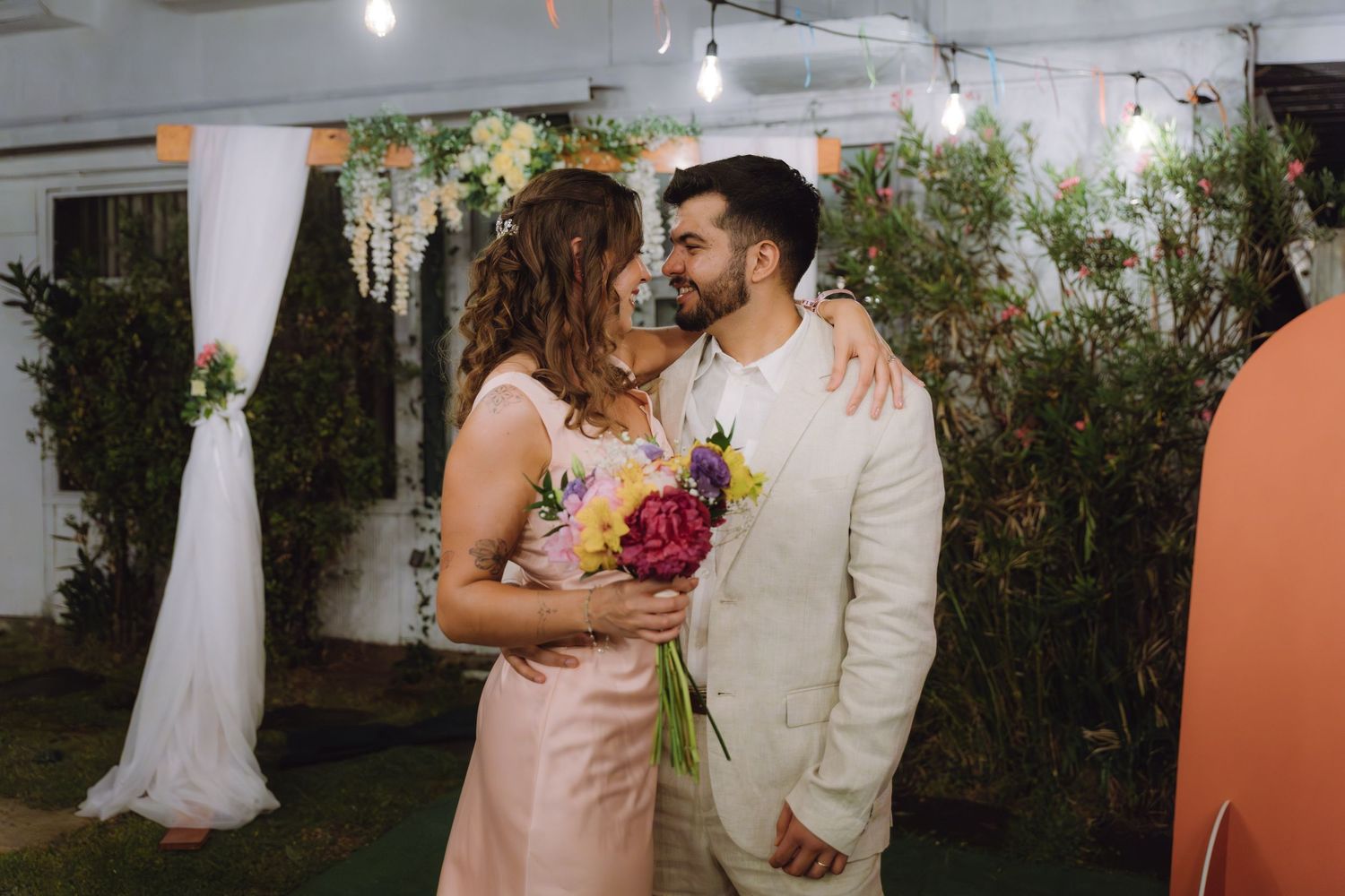 Bride and groom smile at each other holding colorful bouquet at decorated outdoor wedding reception venue.