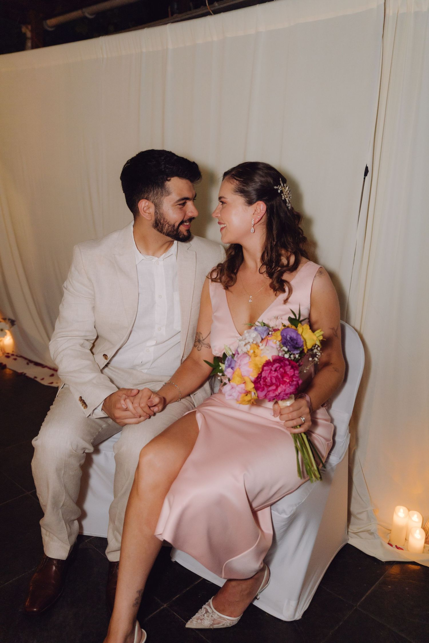 Bride in pink satin dress and groom in white suit sit holding hands with colorful bouquet by candlelight.