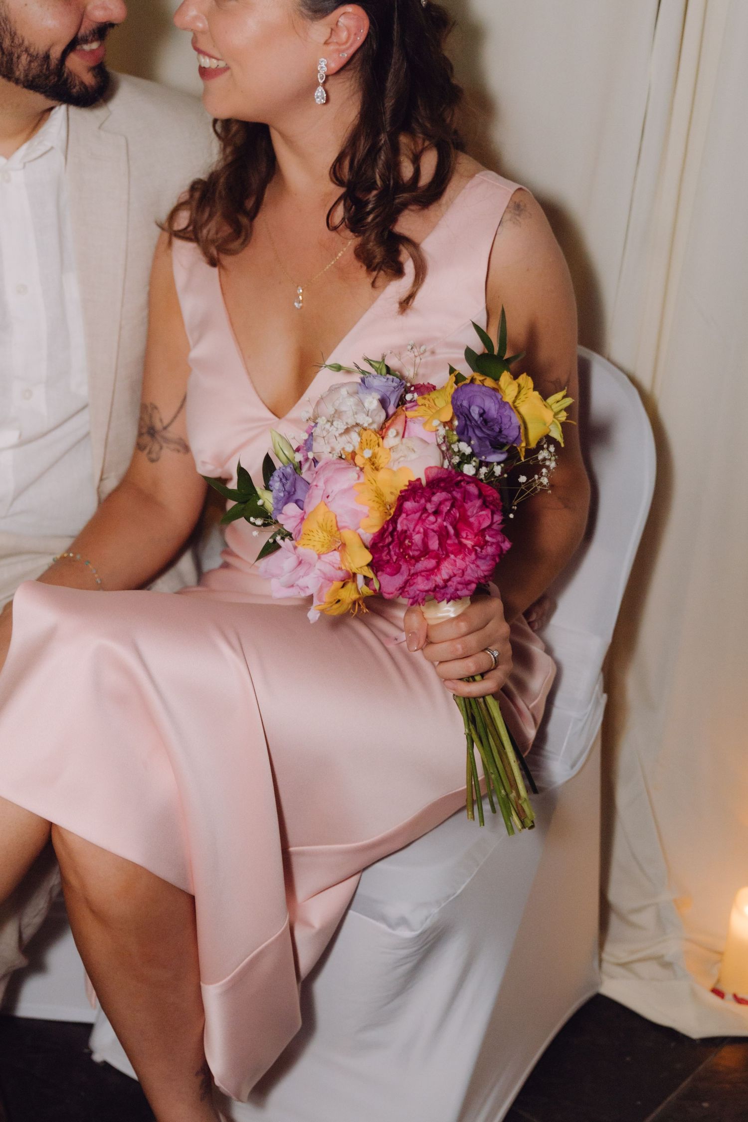 Close-up of bride in pink satin dress holding vibrant bouquet of pink hydrangeas and yellow flowers while seated.