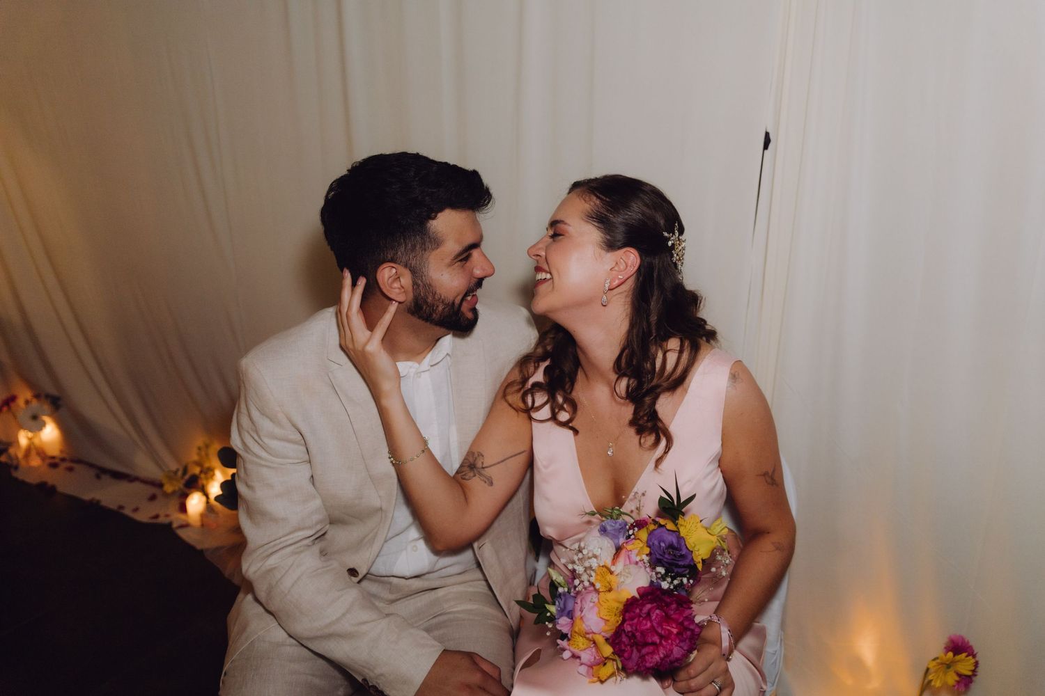 Bride and groom laugh together while seated, she holds colorful bouquet as he touches her face tenderly.