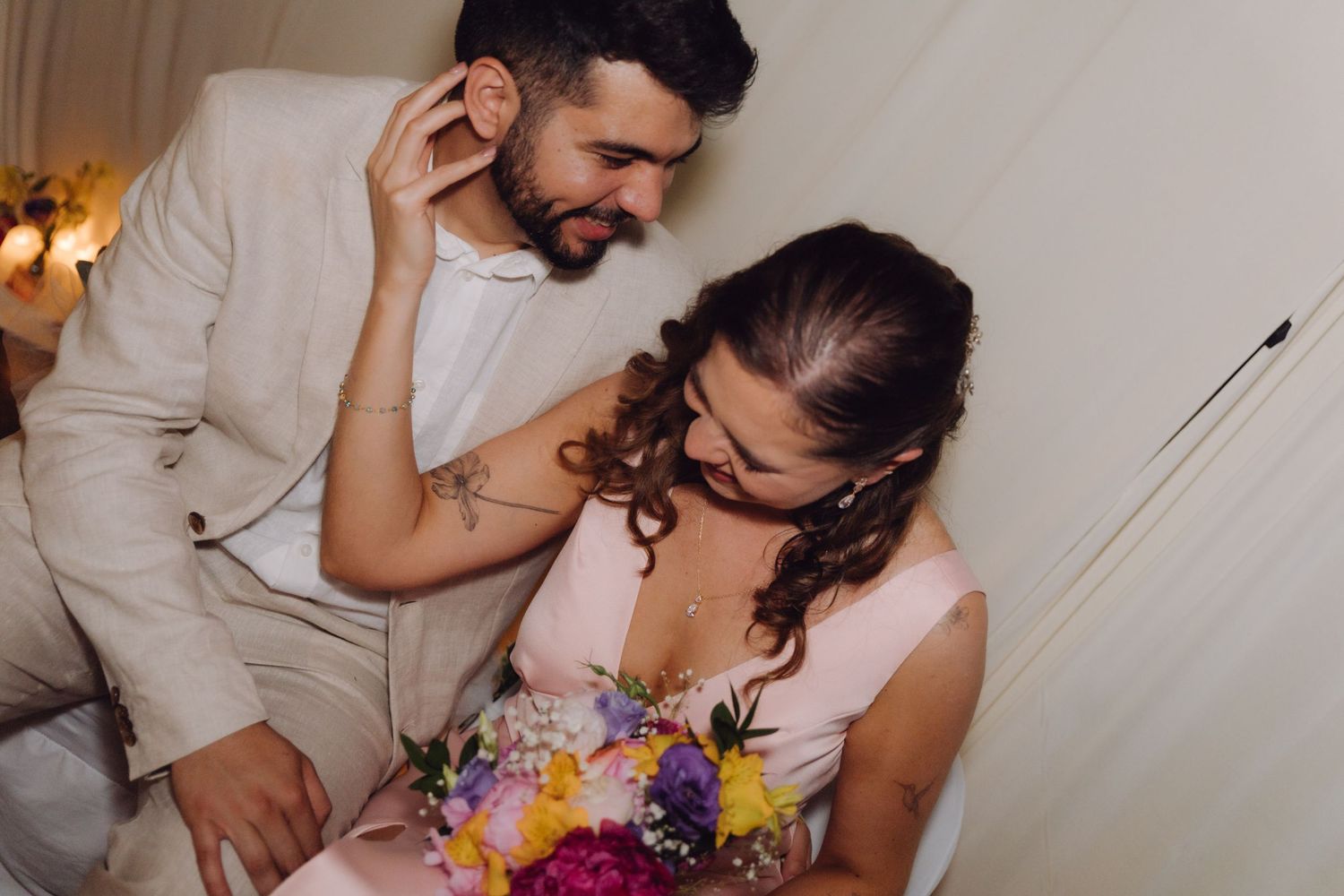 Groom in beige suit tenderly touches brides hair as she looks down holding bright wedding bouquet indoors.