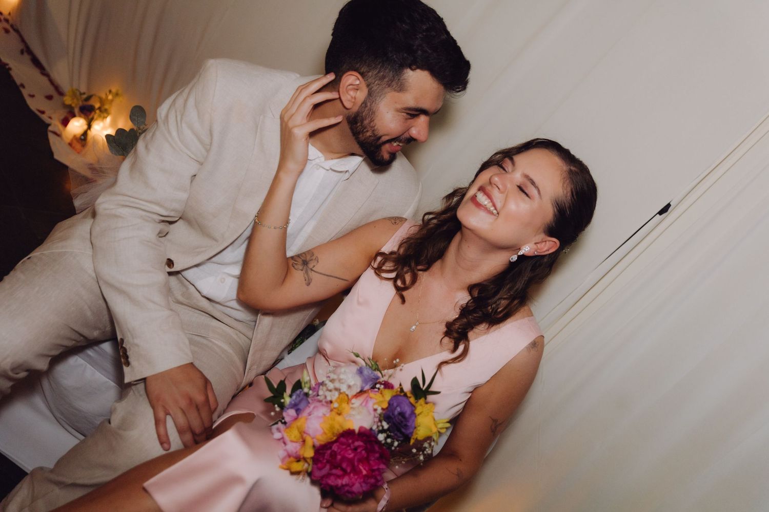 Bride laughs joyfully as groom leans close whispering to her while she holds colorful floral bouquet indoors.