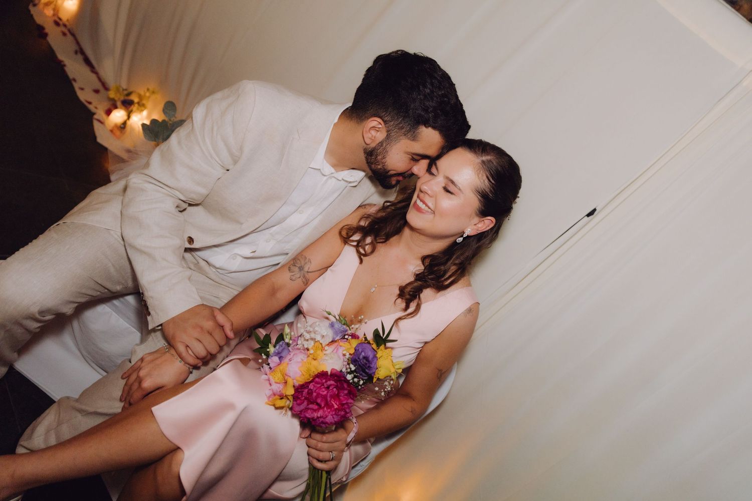 Groom in white linen suit tenderly kissing bride holding vibrant floral bouquet at wedding celebration.
