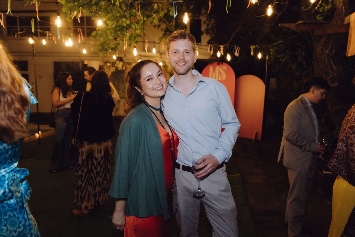 Man and woman smiling together at an outdoor evening party with string lights and guests in background.