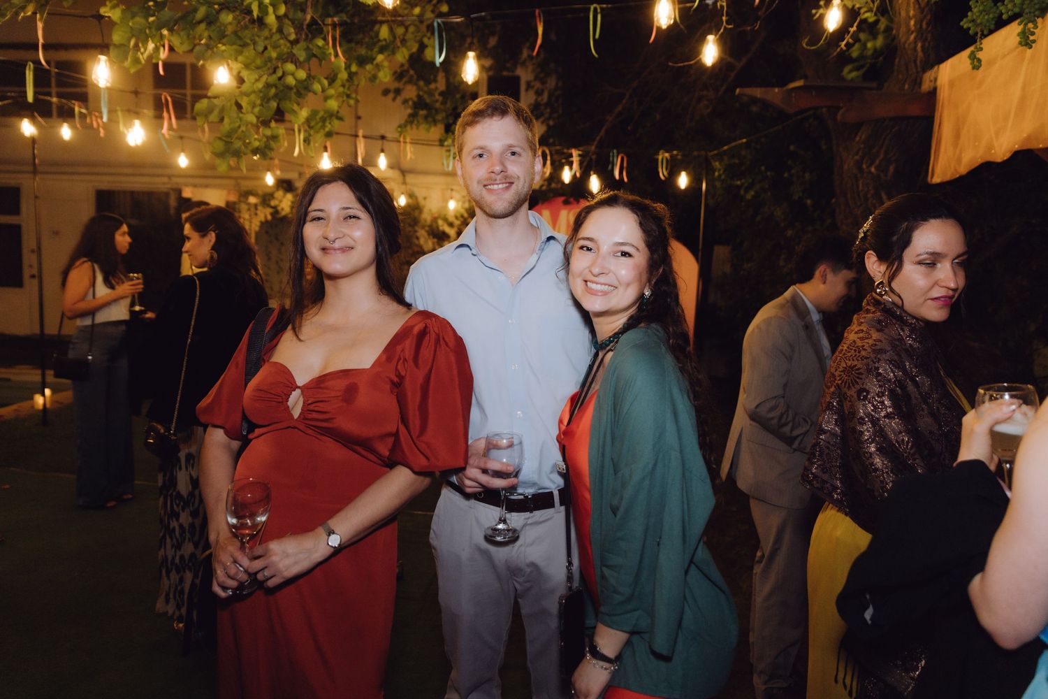 Three guests posing together at an outdoor night event with warm string lights and greenery backdrop.