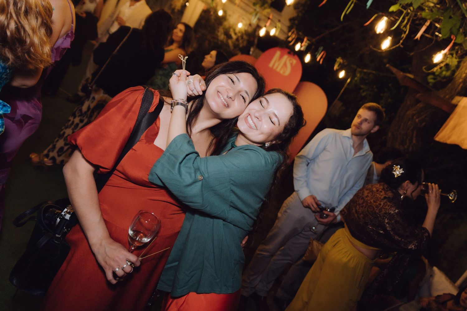 Two women hugging and laughing at an outdoor evening celebration with string lights in background.