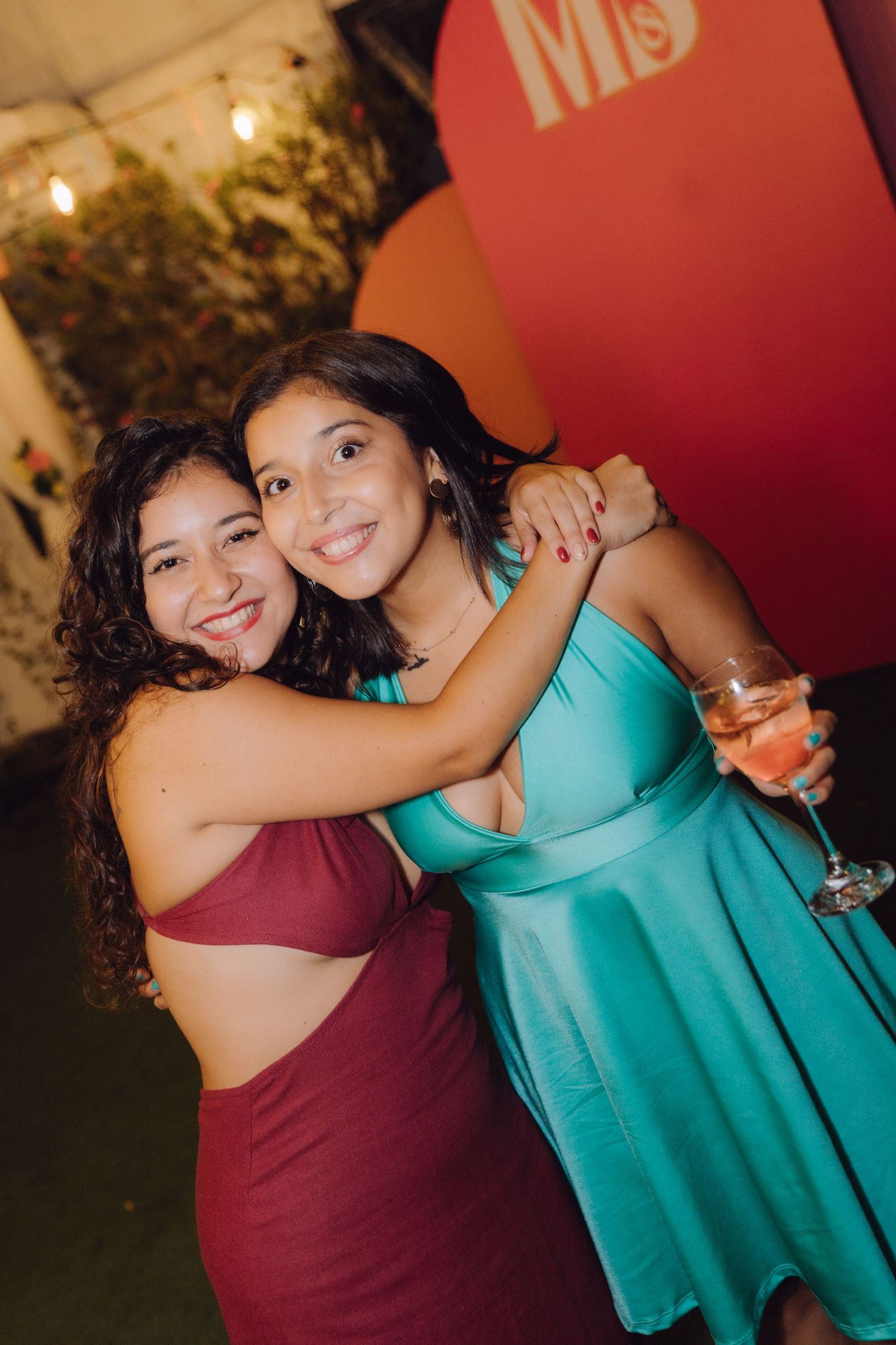 Two young women embracing and smiling at a festive evening party, one holding a pink cocktail glass.