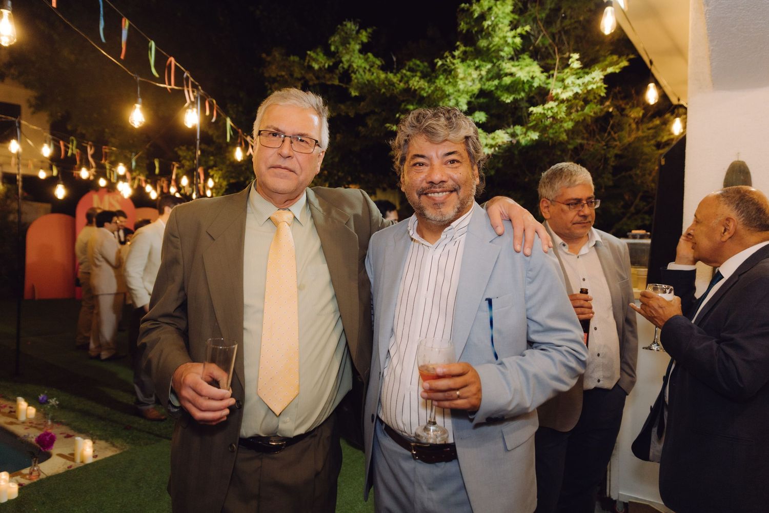Two older gentlemen smiling and holding drinks at an outdoor evening party with string lights.