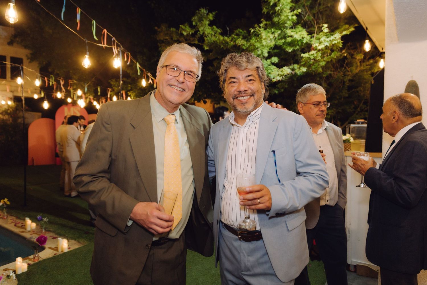 Two men in suits smile and hold drinks at an elegant outdoor evening party with string lights.