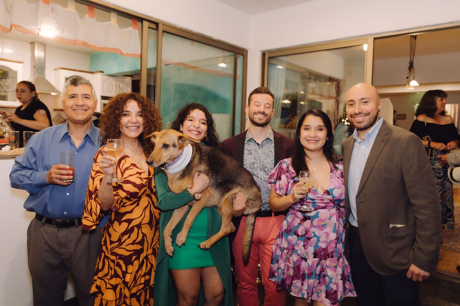 Group of six people and a dog pose together smiling at an indoor evening celebration.