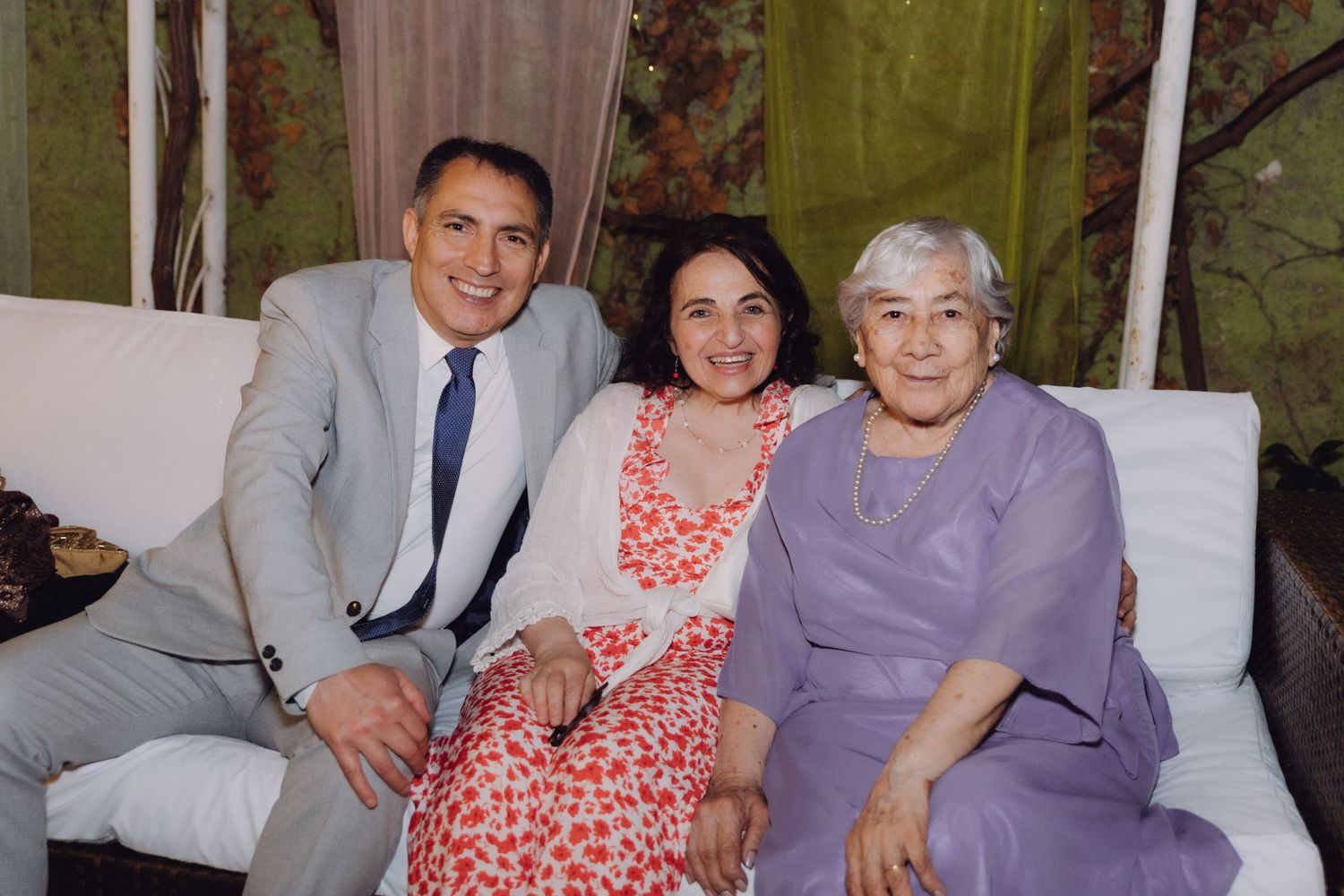 Three people sit together smiling on a white sofa at an intimate indoor gathering.