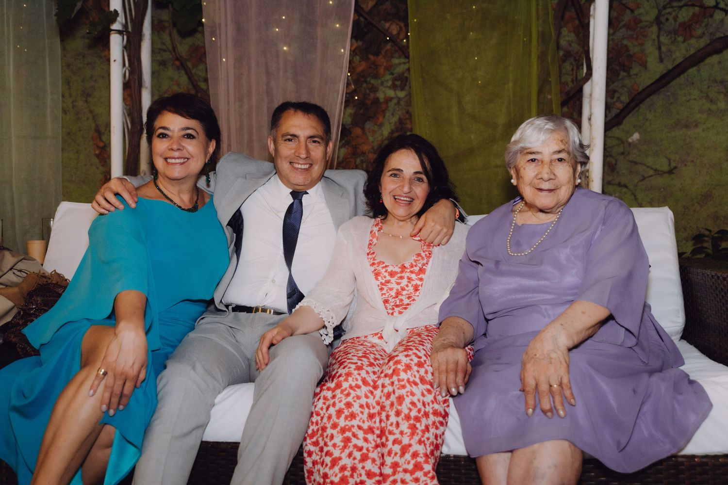 Four people sit closely together on a white sofa, smiling at a social gathering.