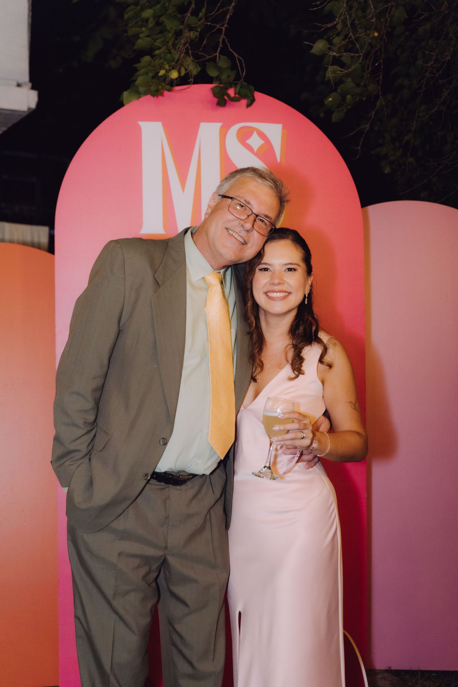 Older man in gray suit and young woman in pink dress pose together at an MS monogram backdrop.