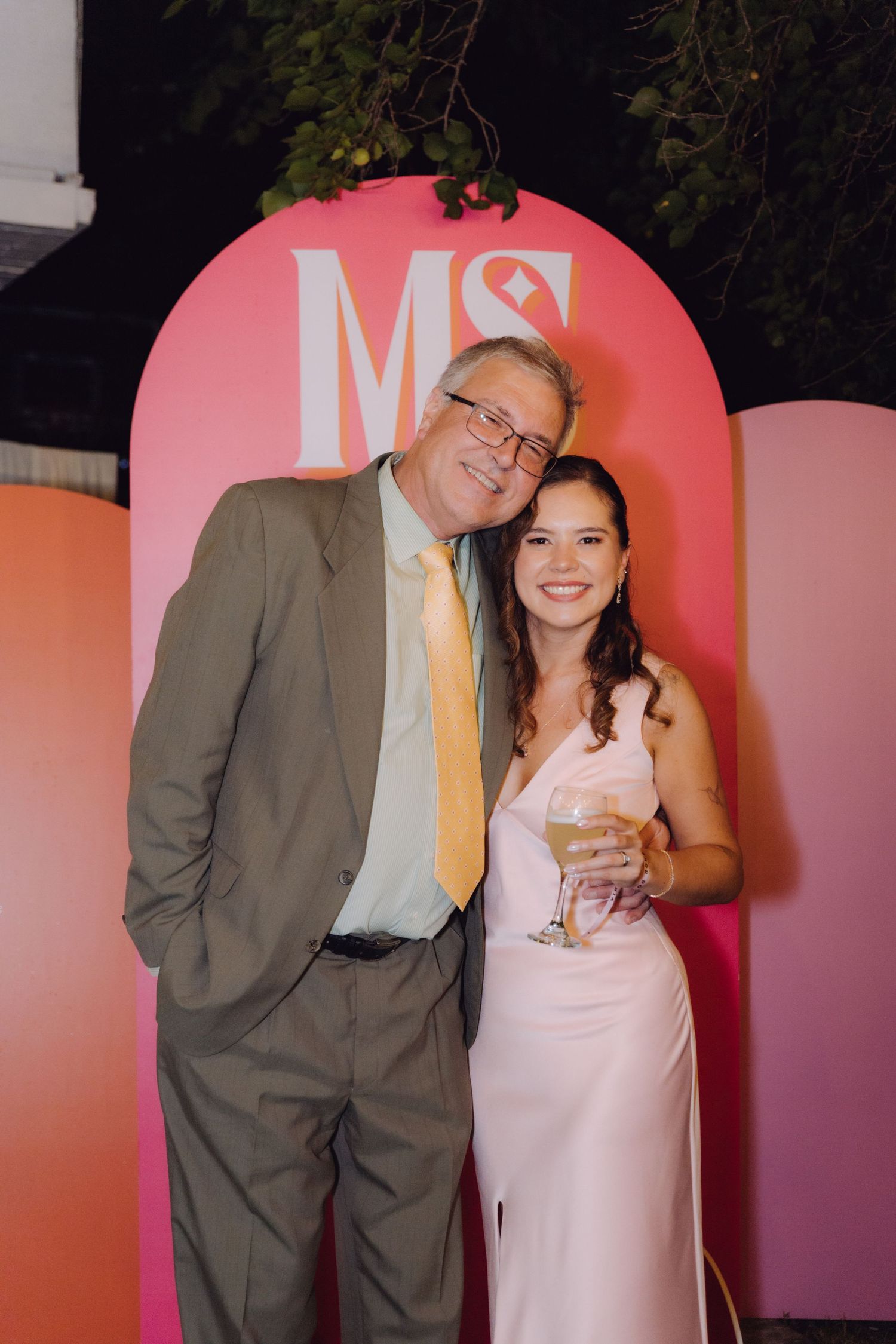 An older man and young woman in a pink dress pose in front of a pink MS monogram backdrop.