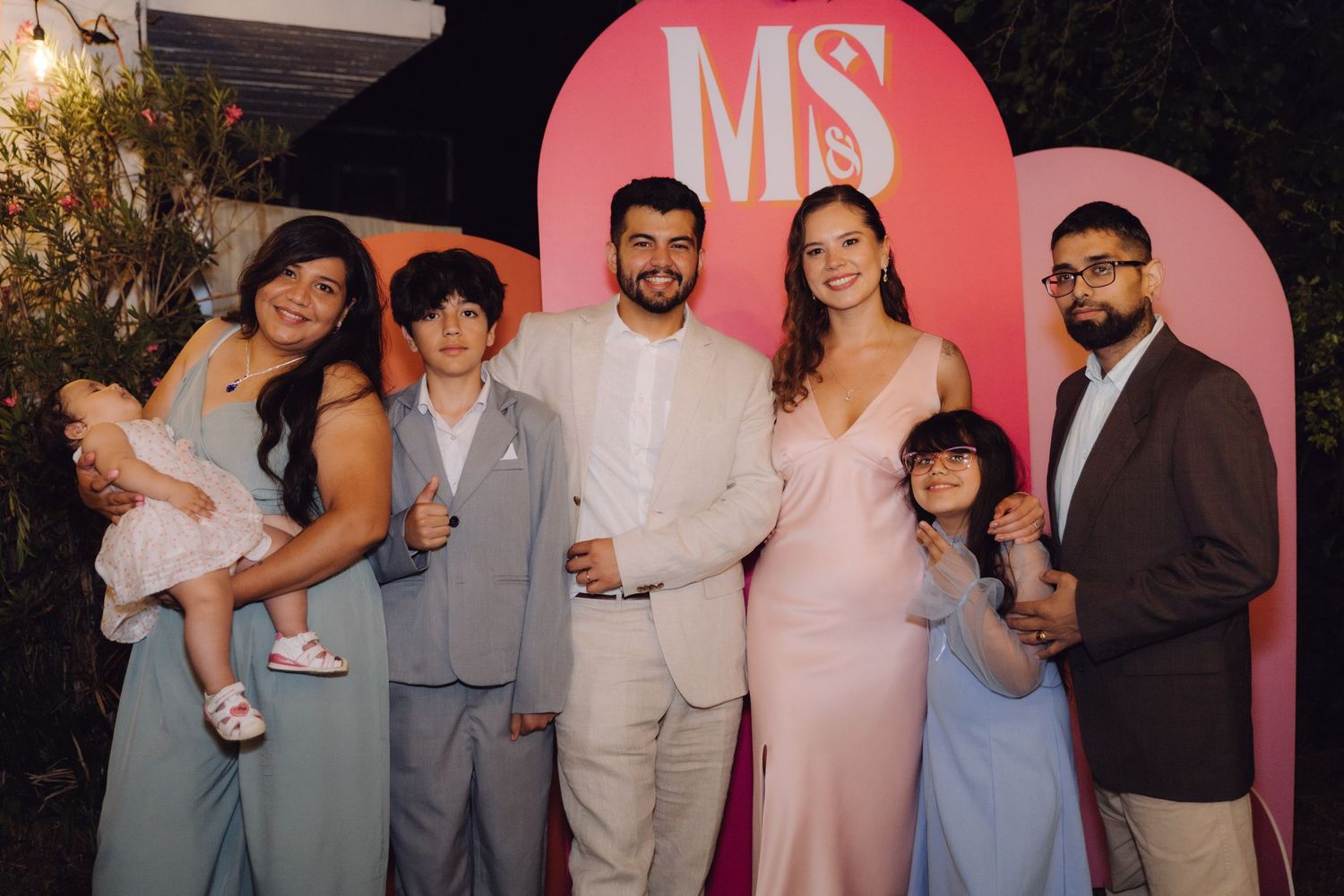 Family group of six adults and children pose together in front of pink MS monogram wedding backdrop.