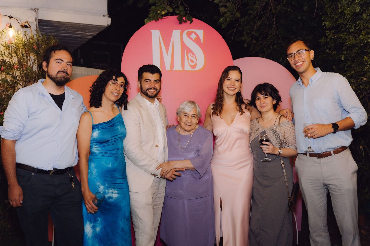 Group of seven people including elderly woman pose together at pink MS monogram event backdrop.