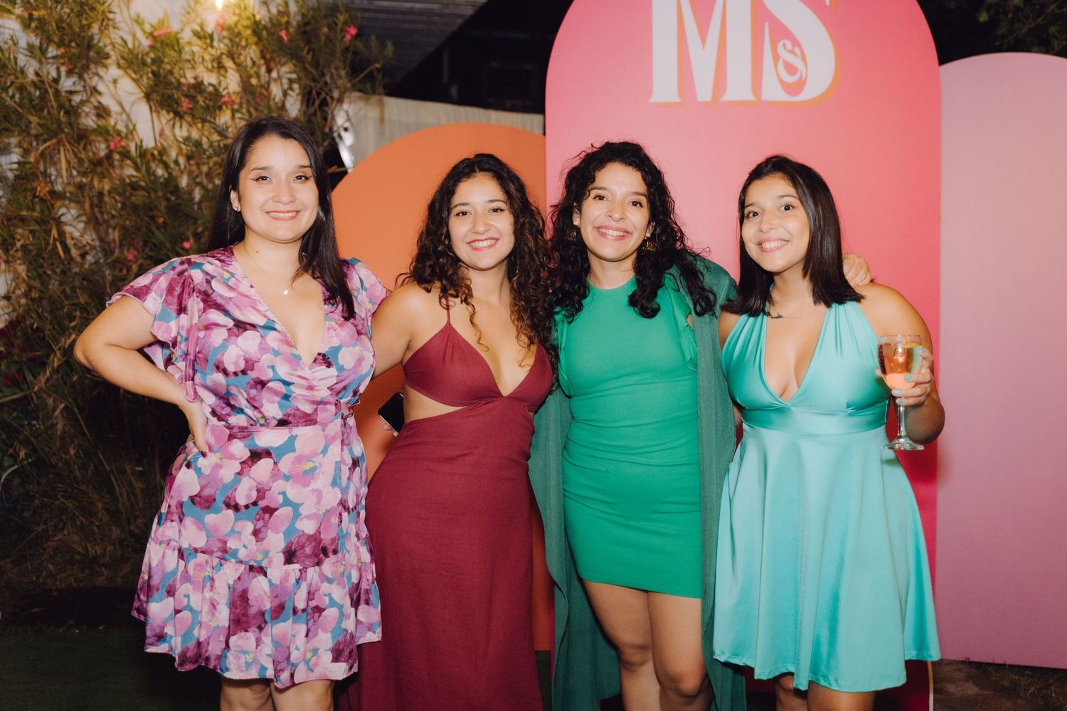 Four young women in colorful dresses smile together in front of pink MS monogram event backdrop.