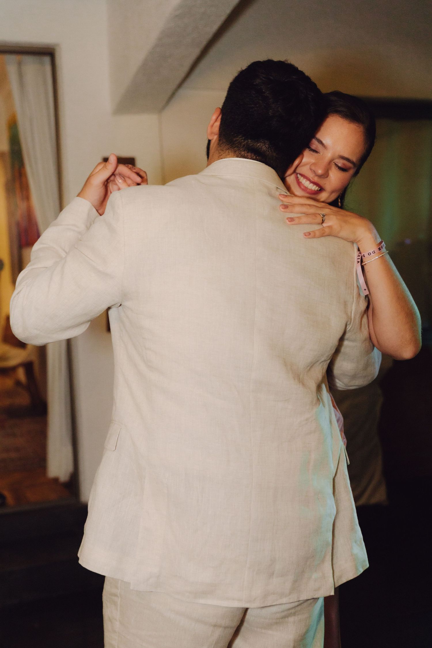 Smiling bride in pink dress embraces groom in beige suit during their first wedding dance.