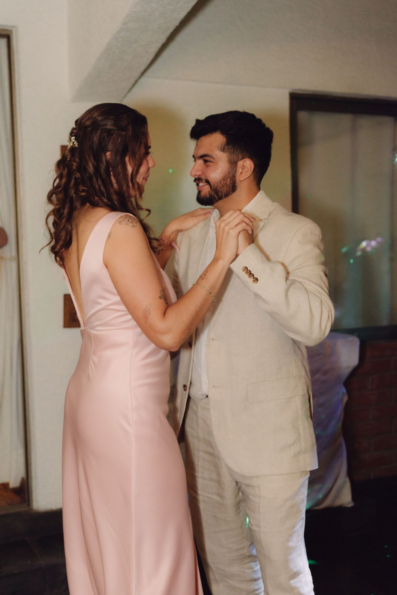 Bride in pink dress adjusts groom's beige suit collar as they smile at each other.