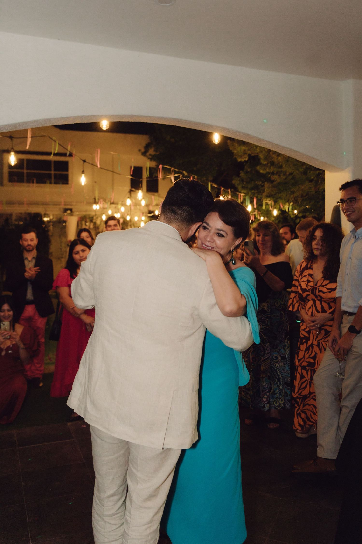 A smiling woman in a teal dress shares a tender dance with a man in a beige suit at an elegant evening wedding reception.