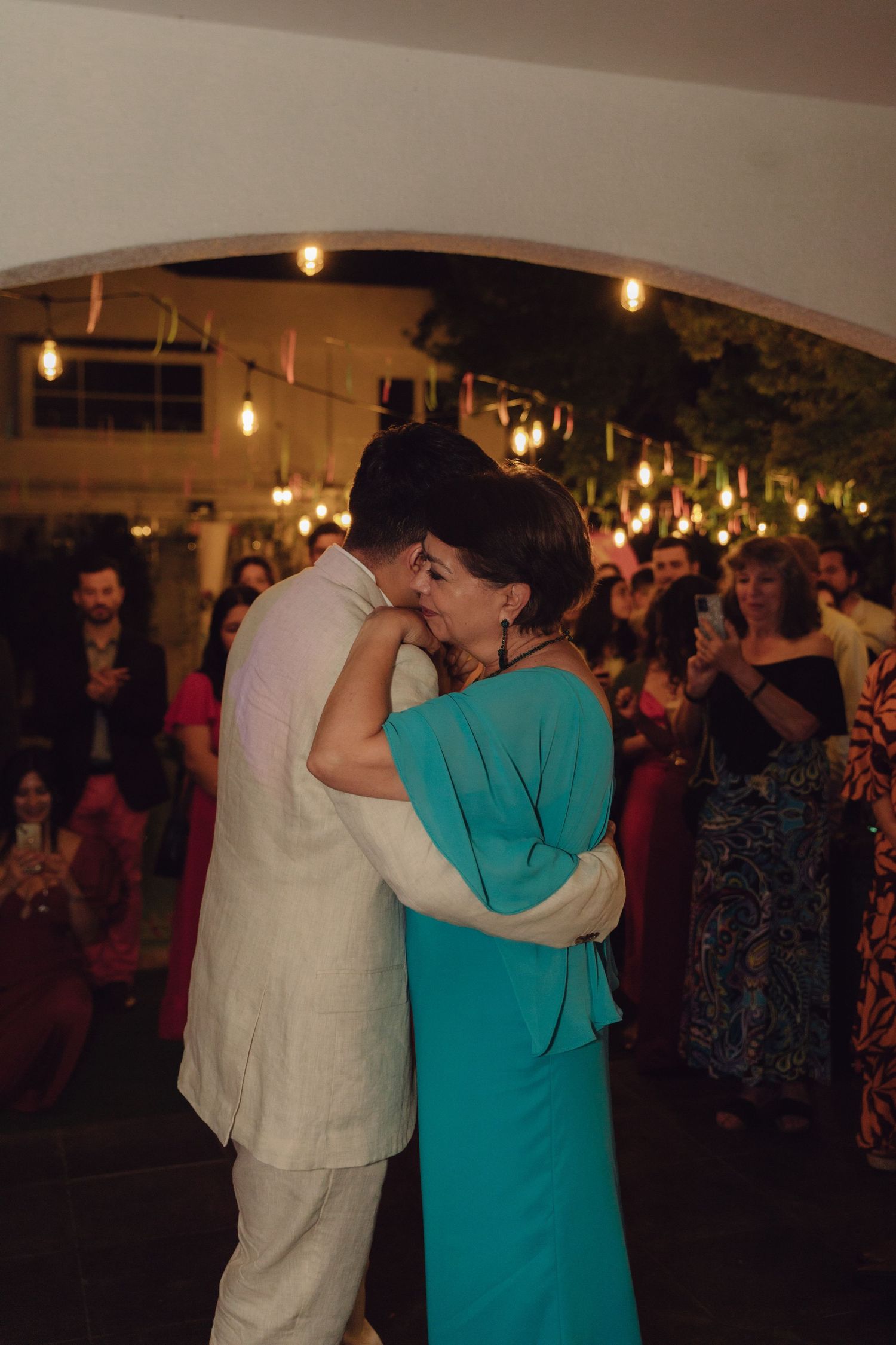 A couple shares an emotional embrace while dancing at a warmly lit wedding reception with guests watching in the background.