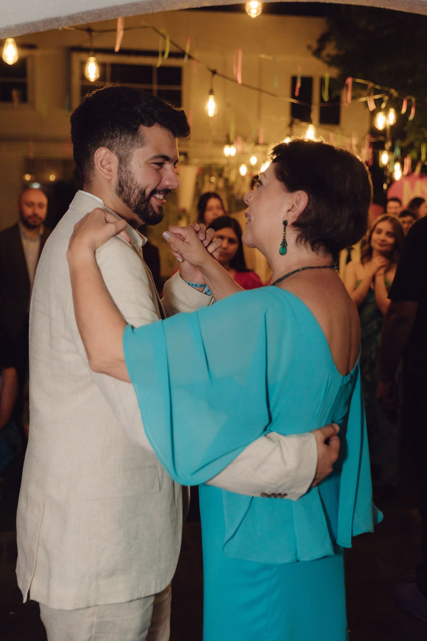 A young man in a beige suit and a woman in a teal off-shoulder dress share a joyful mother-son dance at a wedding.