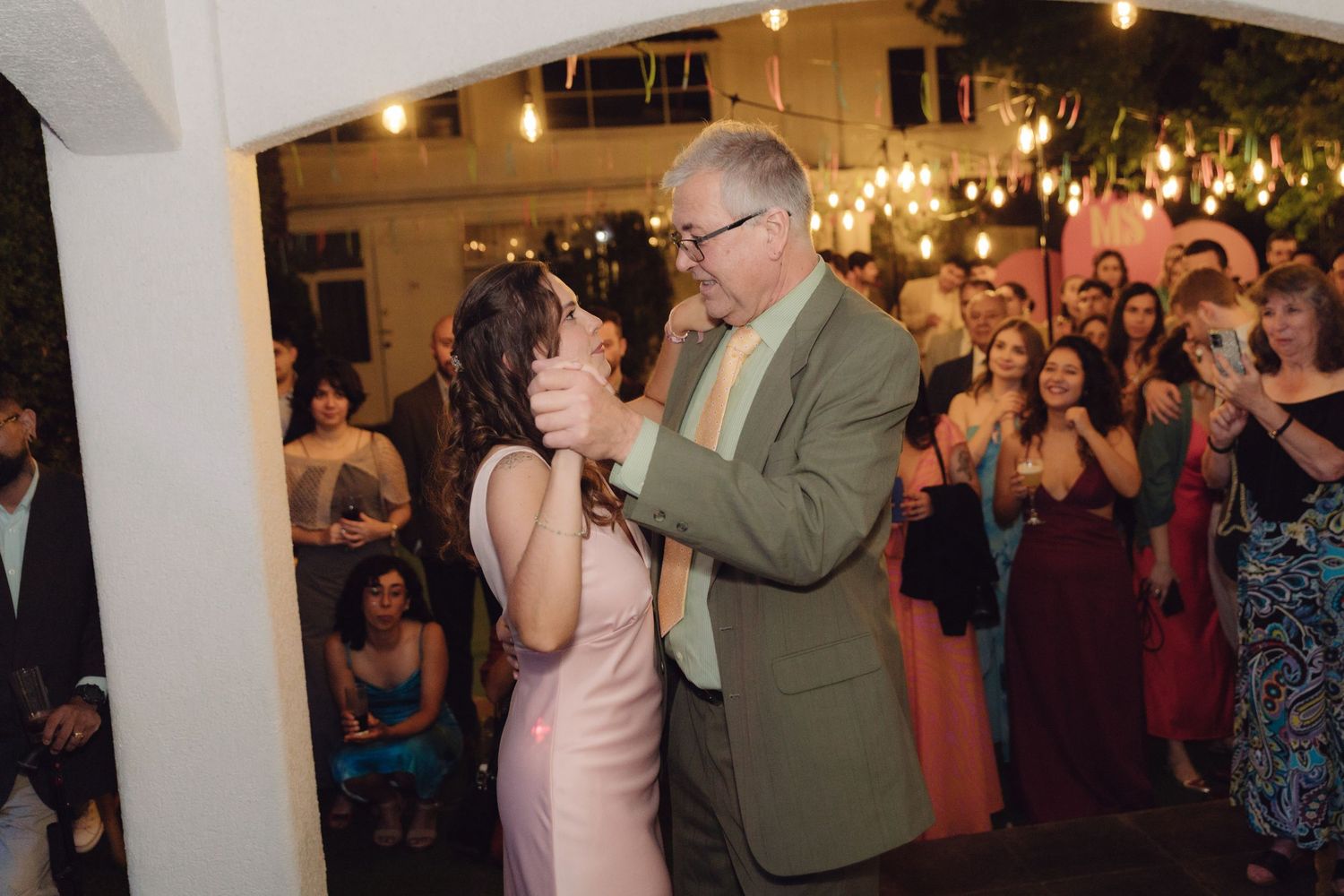 A woman in a pink dress dances with an older man in a green suit under string lights at a wedding reception.