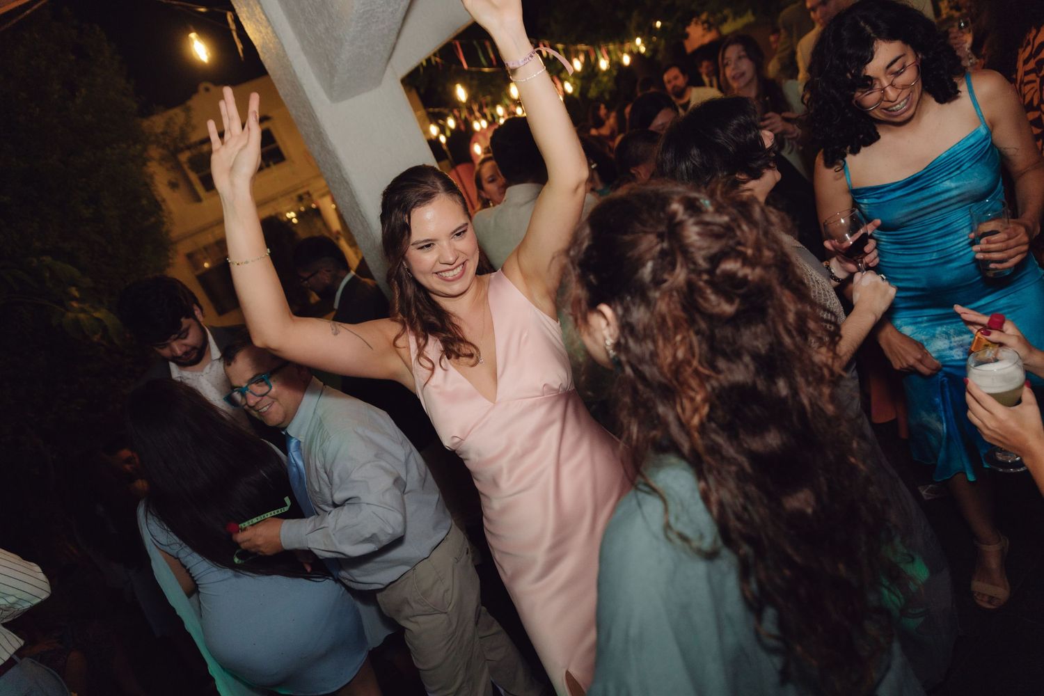 A bridesmaid in a pink dress dances with arms raised joyfully on a crowded wedding reception dance floor at night.