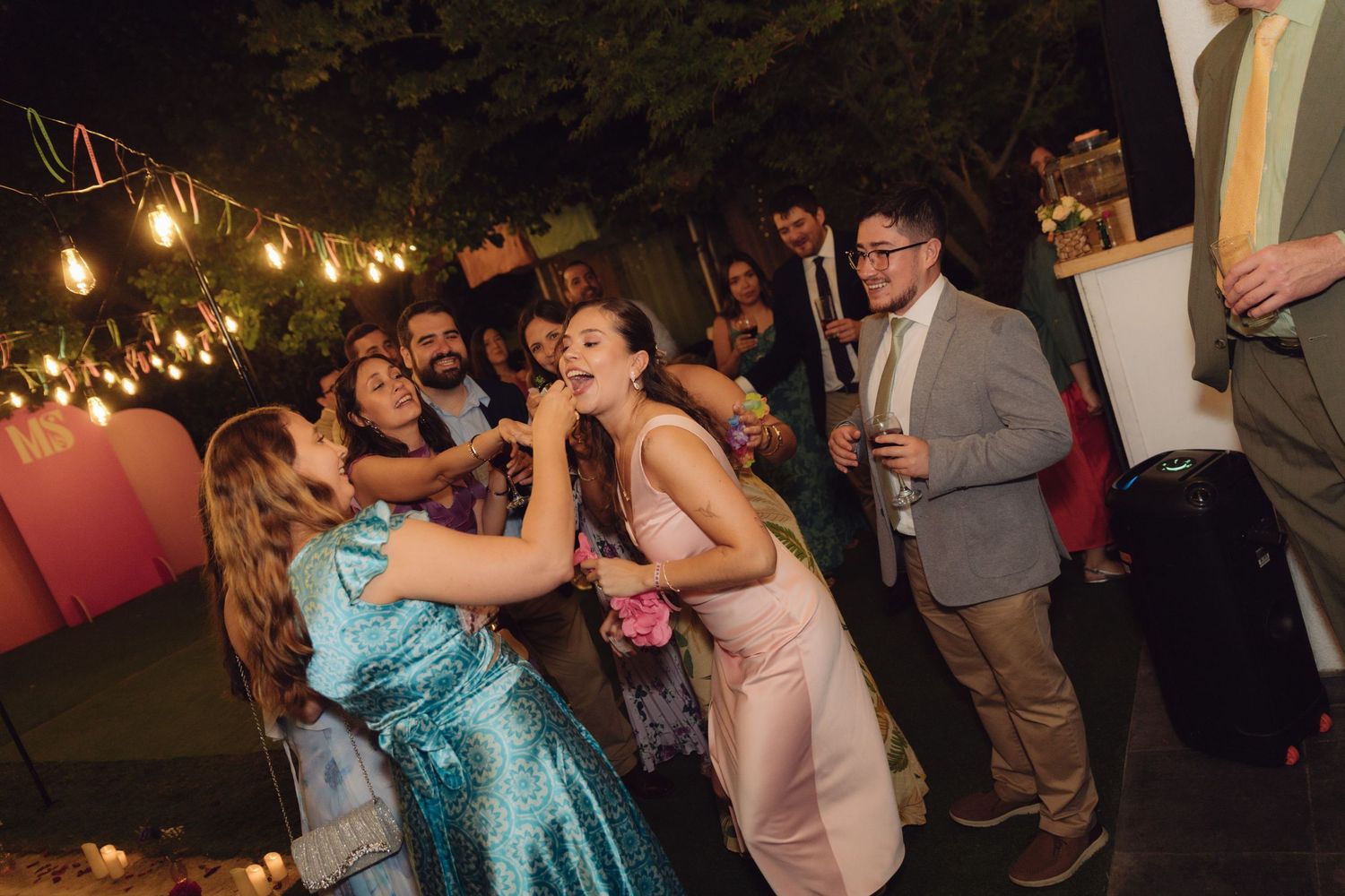 Wedding guests laugh and dance outdoors at night under string lights during a festive wedding reception celebration.