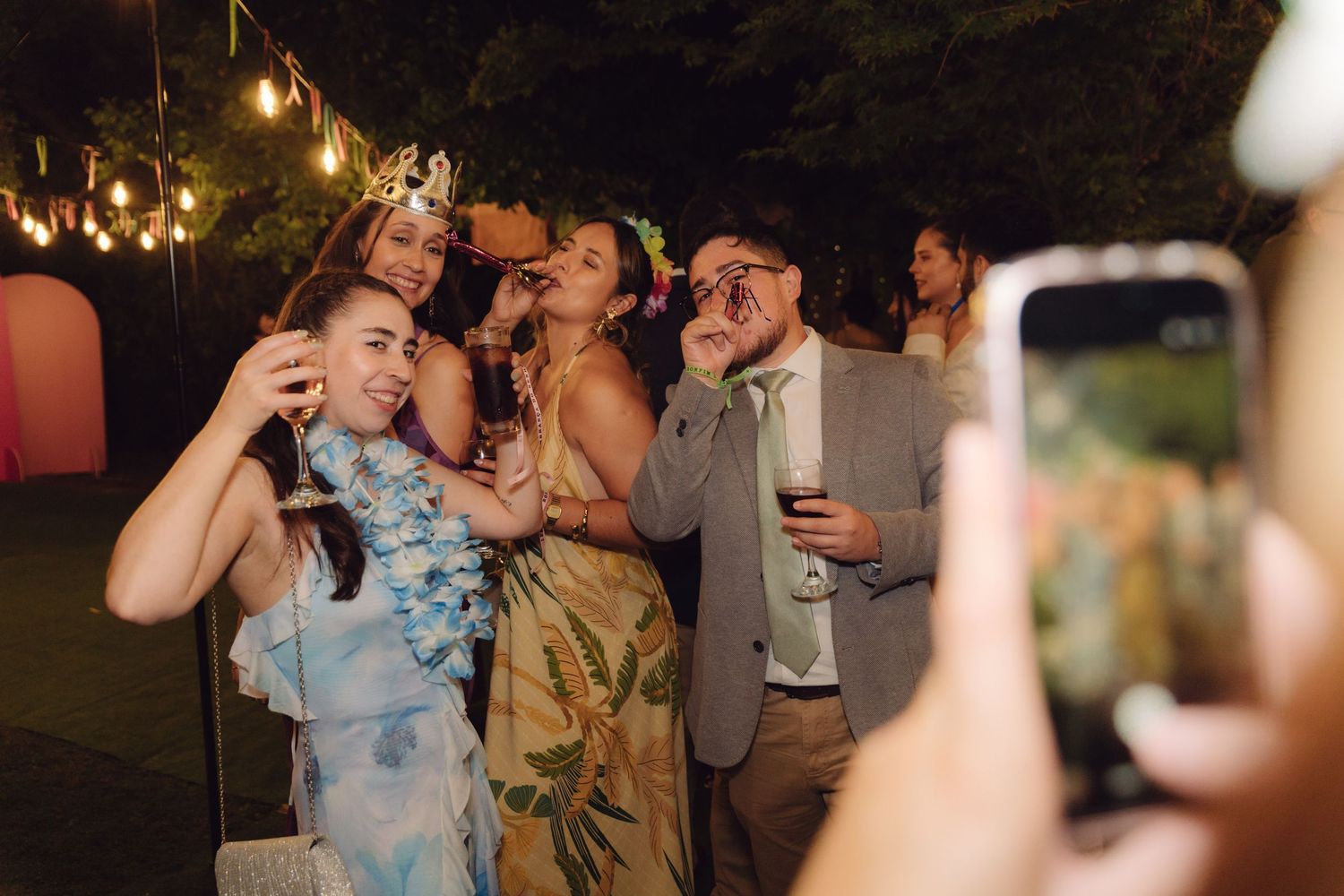 A group of wedding guests pose playfully for a smartphone photo outdoors at night during a wedding reception.
