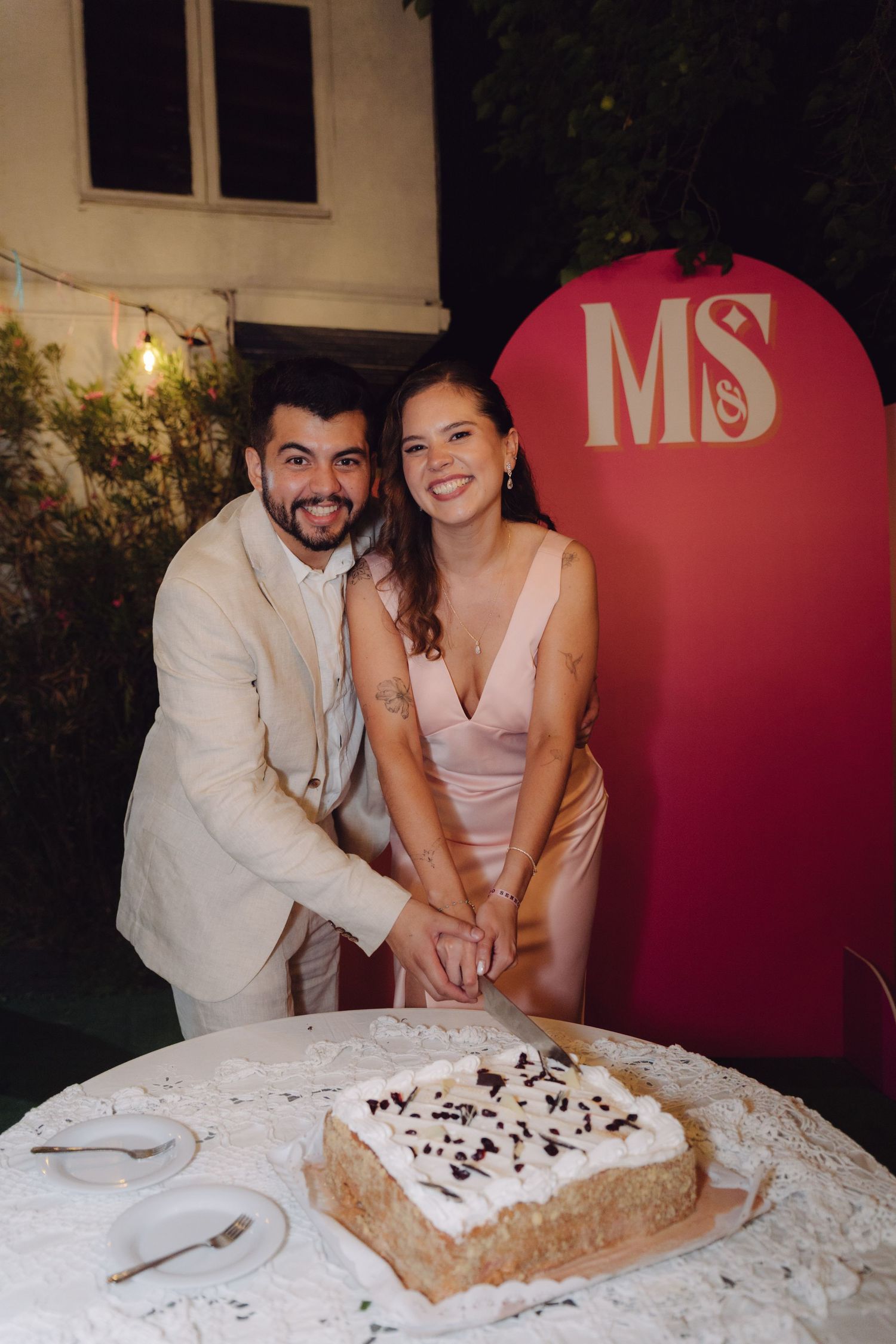 Couple smiling while cutting their wedding cake outdoors at night with MS monogram backdrop.