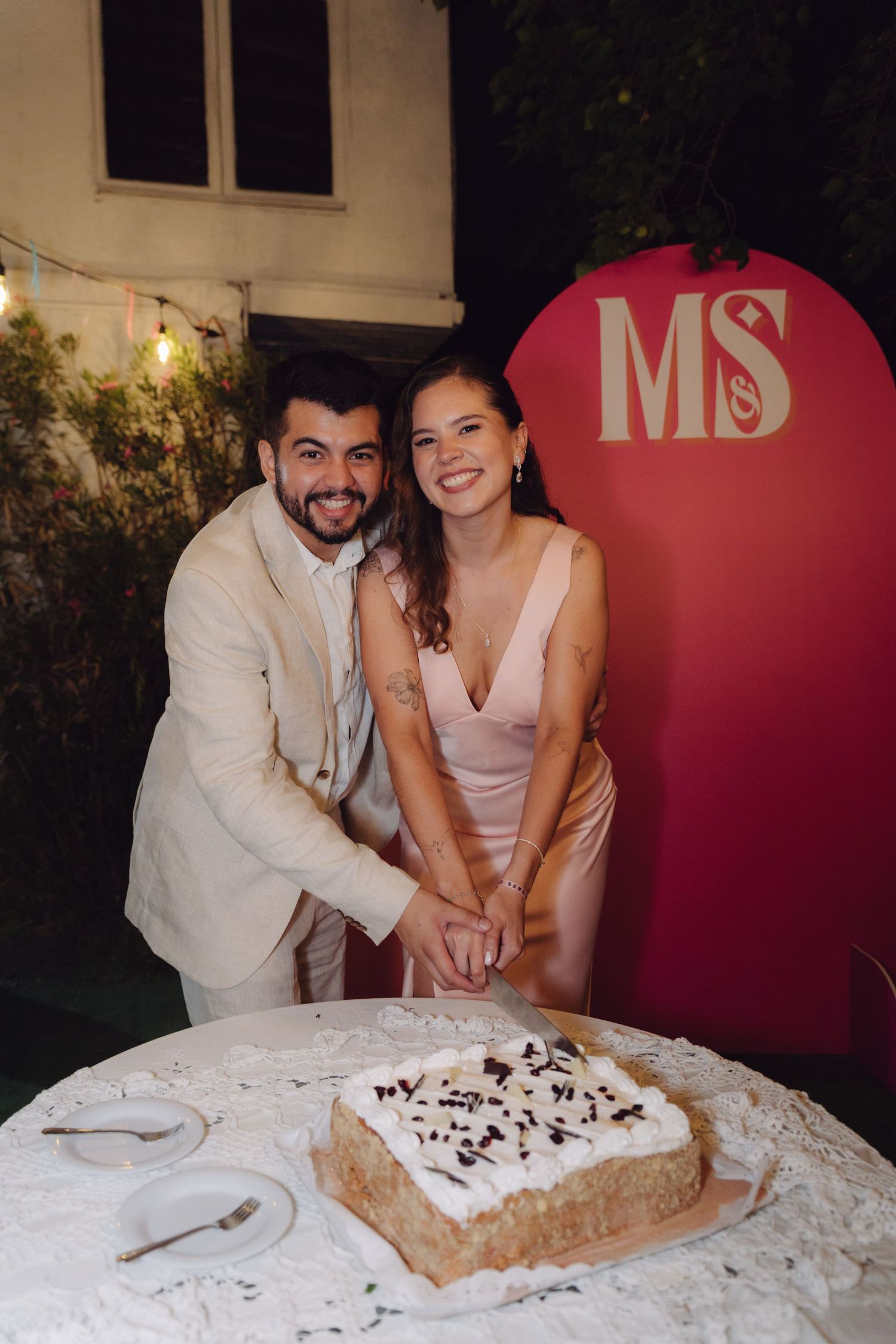 Bride and groom posing together cutting wedding cake with pink MS monogram sign behind them.