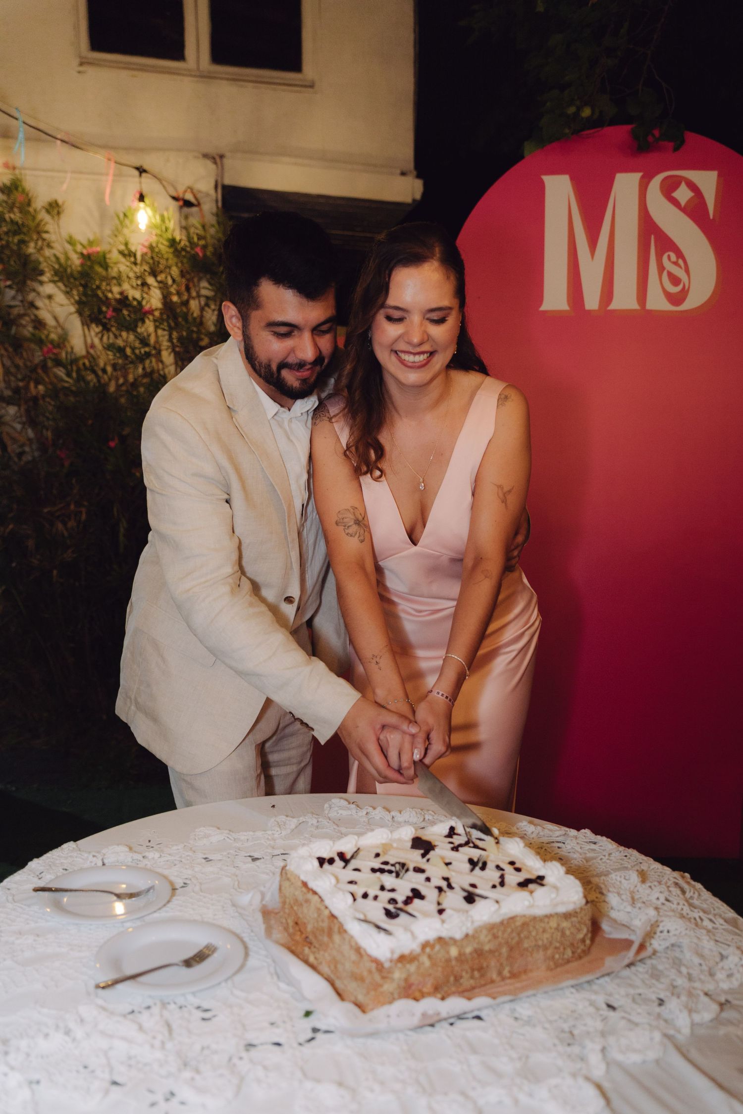 Happy couple cutting wedding cake together outdoors at evening reception with decorative backdrop.