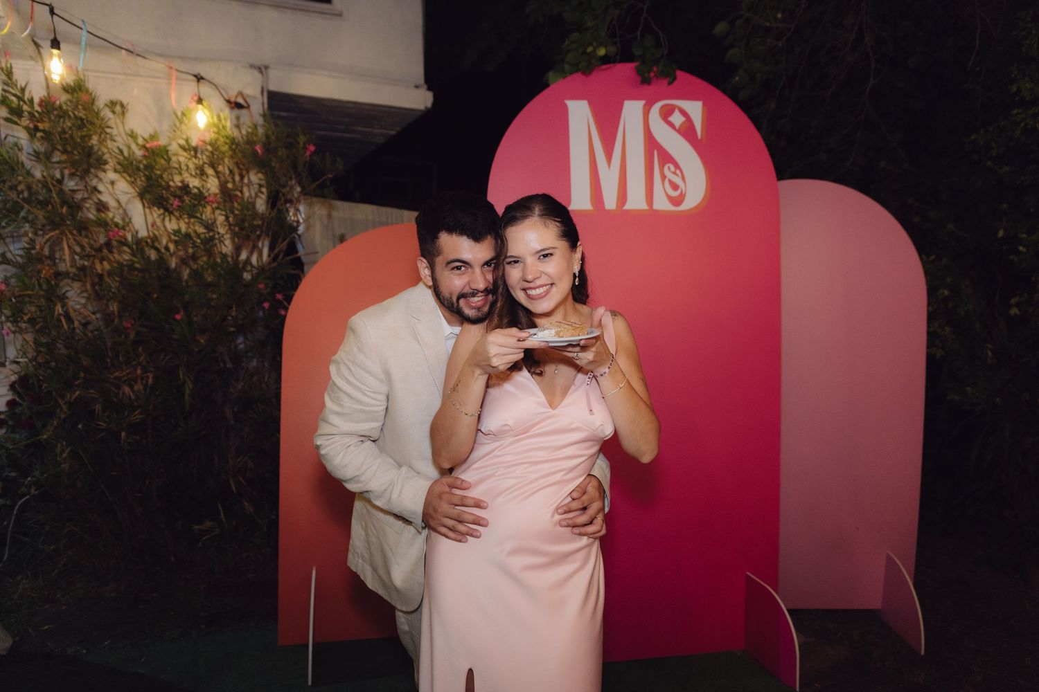 Smiling couple posing together holding cake slice in front of pink MS wedding photo backdrop.