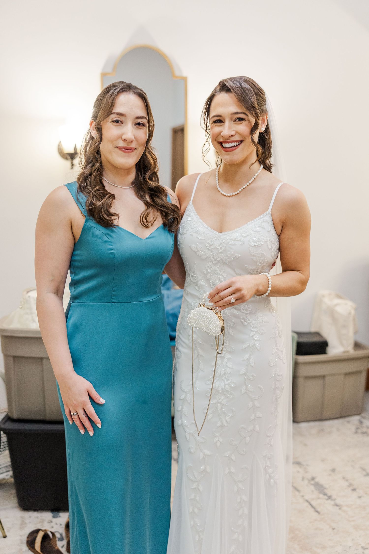 Bride in white lace gown and bridesmaid in teal dress smiling together before wedding ceremony.