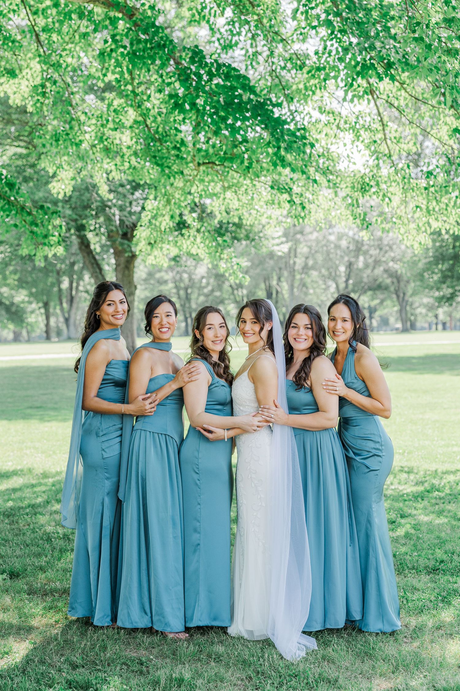 Bride in white gown poses with five bridesmaids in dusty blue dresses outdoors under lush green trees.