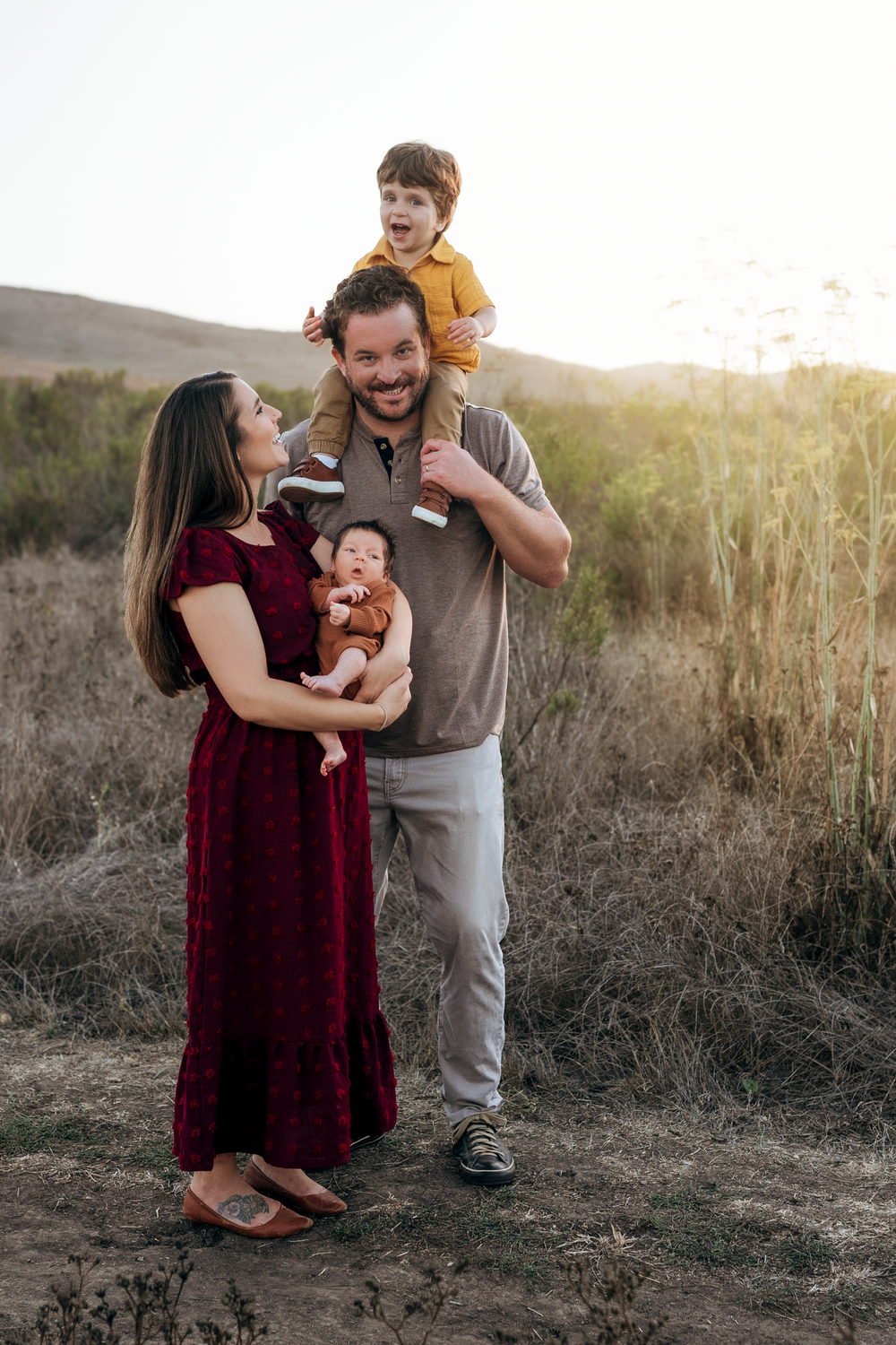 Family of four posing outdoors at golden hour, mother in burgundy dress holding infant, father with toddler on shoulders.