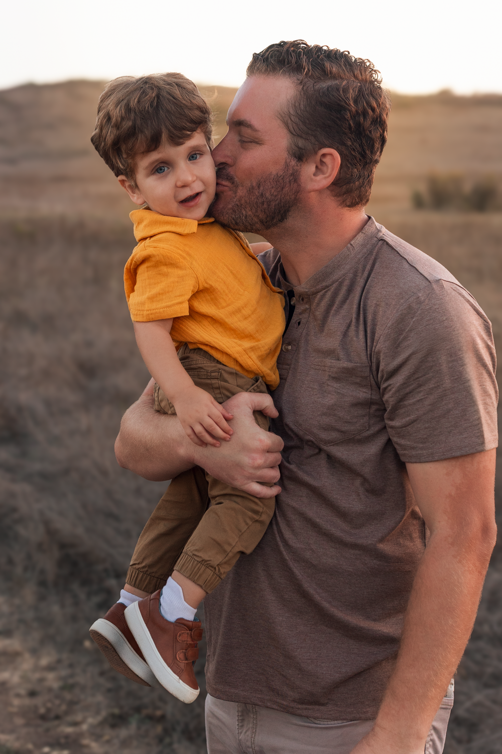 A father kisses his young son on the cheek while holding him in a golden field at sunset.