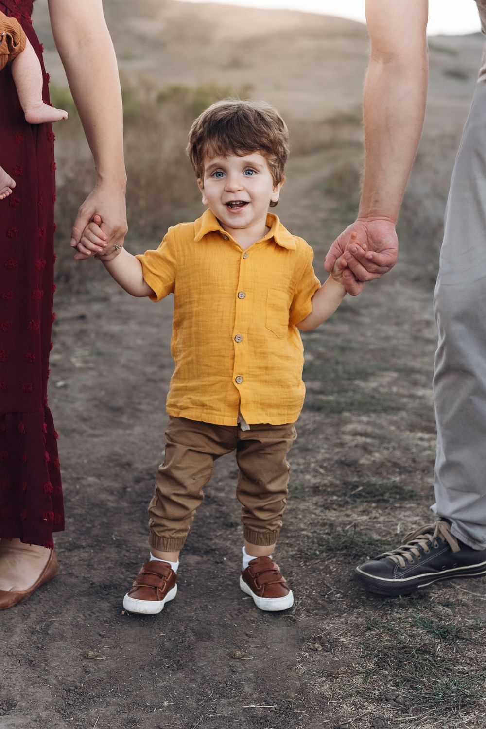 Toddler boy in yellow shirt and brown pants holds parents hands outdoors on a dirt path.
