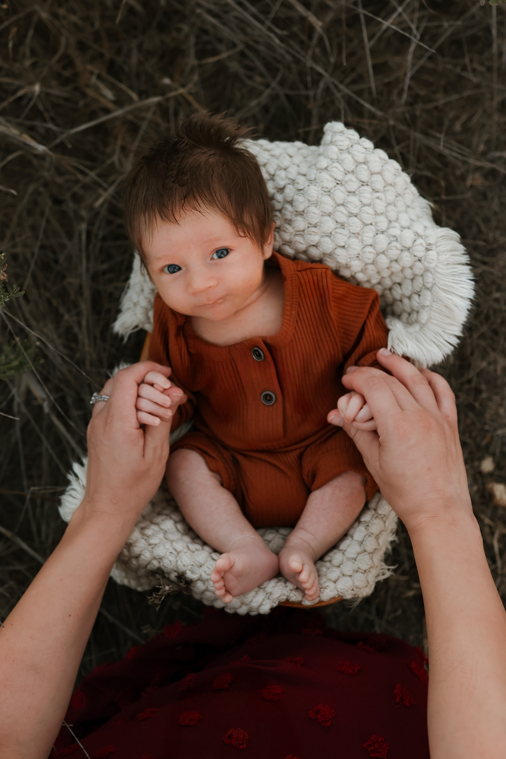 A newborn baby in a rust romper rests on a white knit blanket outdoors, cradled by gentle hands.
