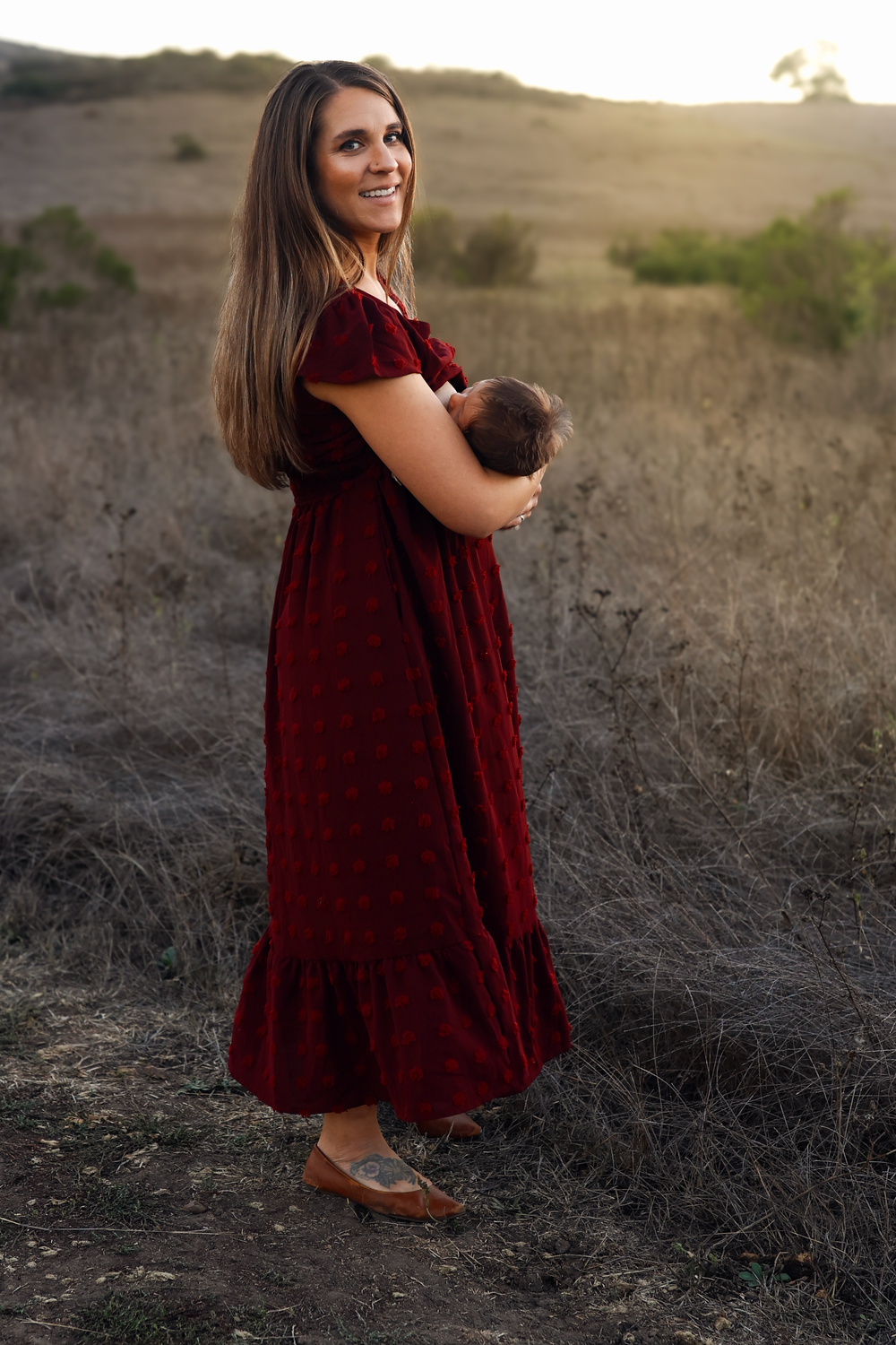 A smiling woman in a red dress holds a newborn baby in a sunlit field at golden hour.