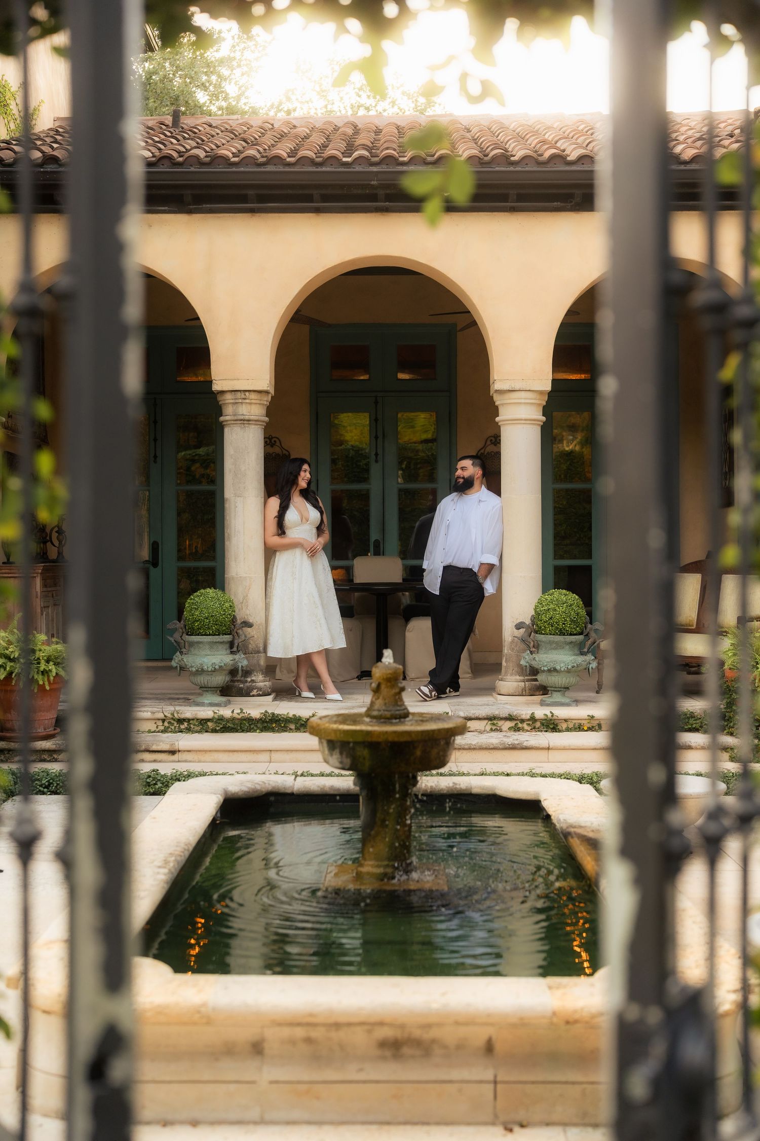 Couple standing by a stone fountain in an elegant courtyard with arched colonnade and lush greenery.
