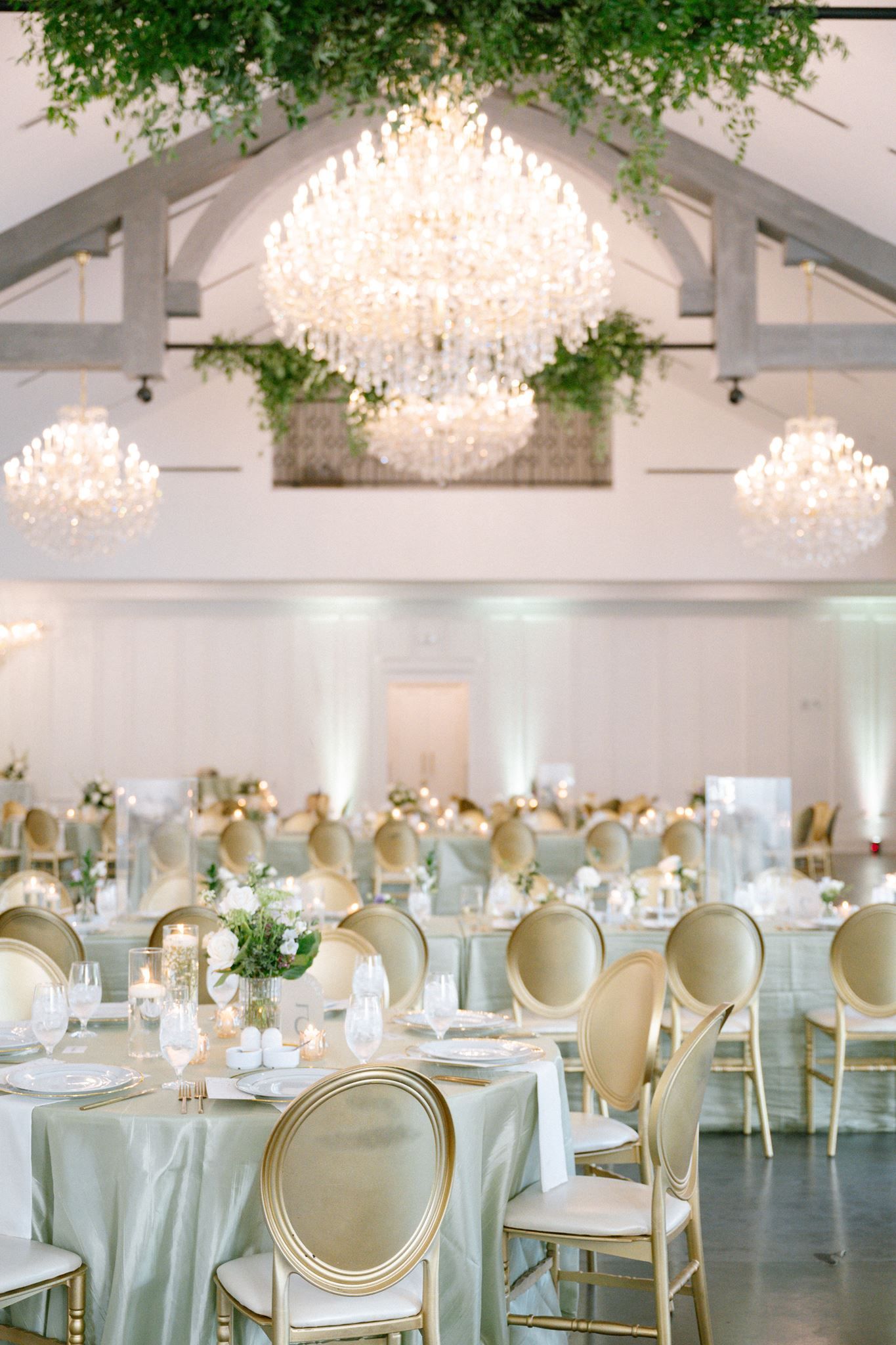 Elegant wedding reception hall with crystal chandeliers, gold chairs, sage green linens, and lush greenery in a bright white venue.