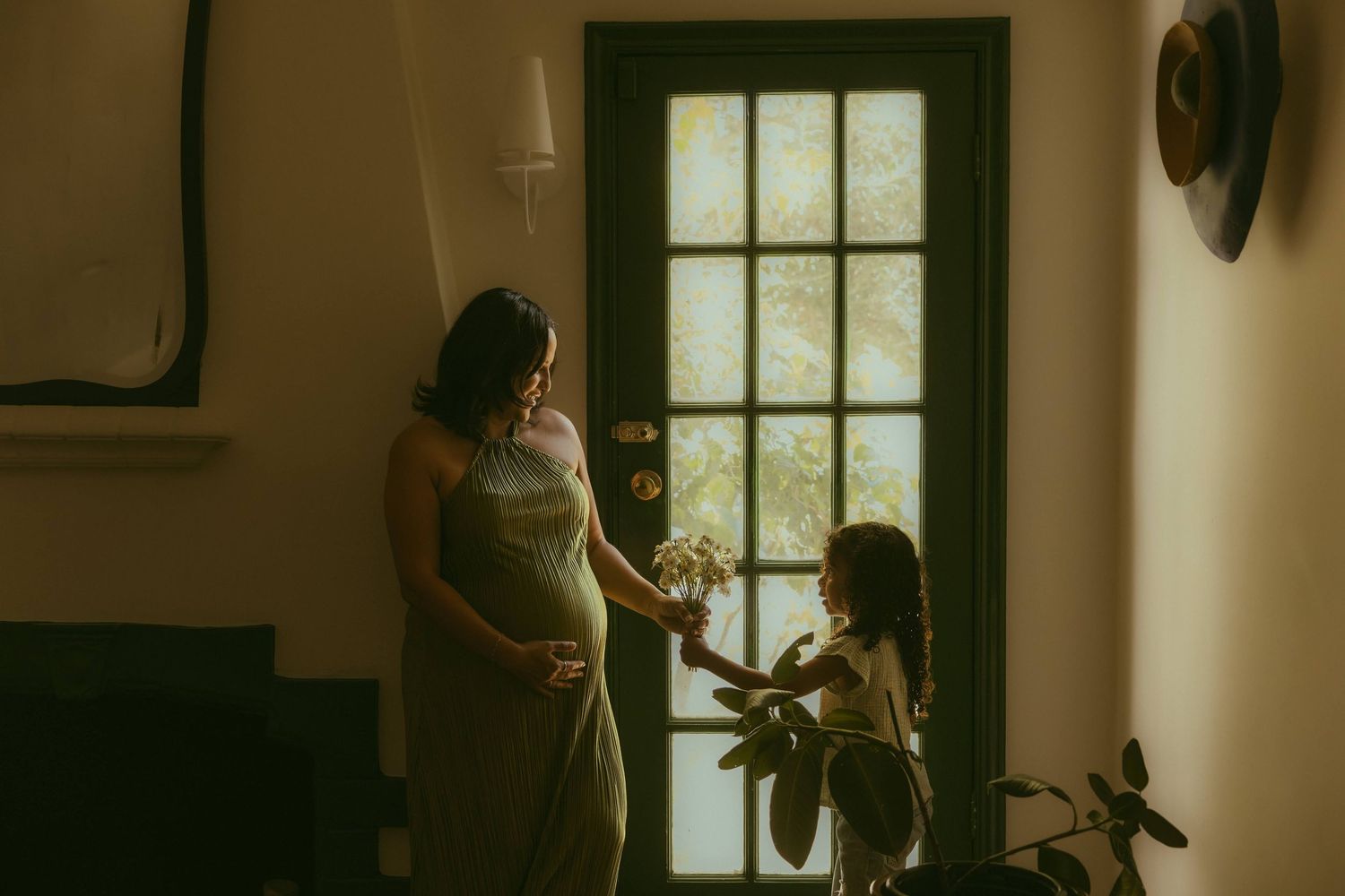 Pregnant woman in olive dress receiving flowers from her daughter during their family photo session in warm natural light.