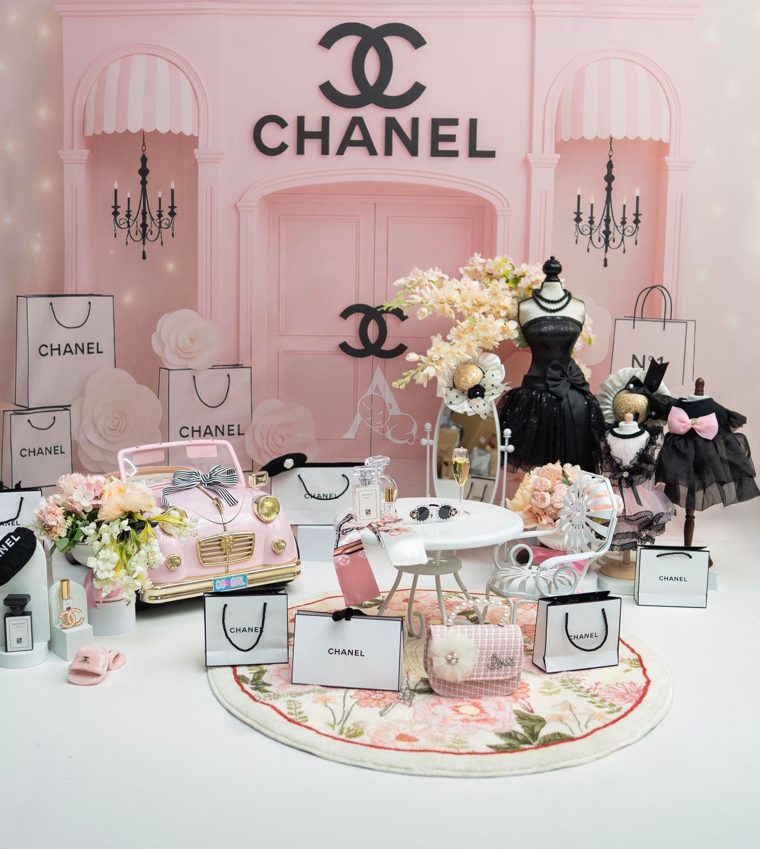 Chanel-themed pink display with shopping bags, flowers, toy car, and black dress mannequin in elegant boutique setting.